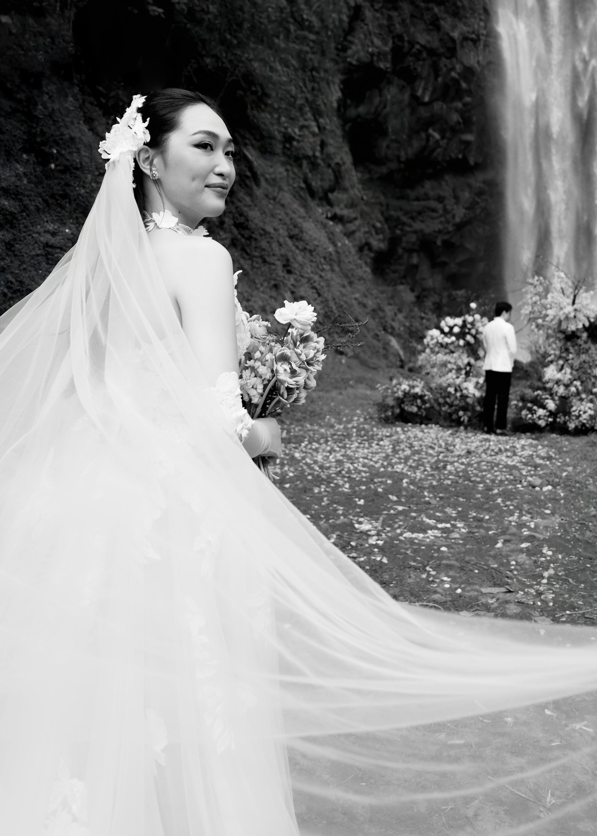 Elegant bride in lace gown with long veil holding bouquet near waterfall in black and white.