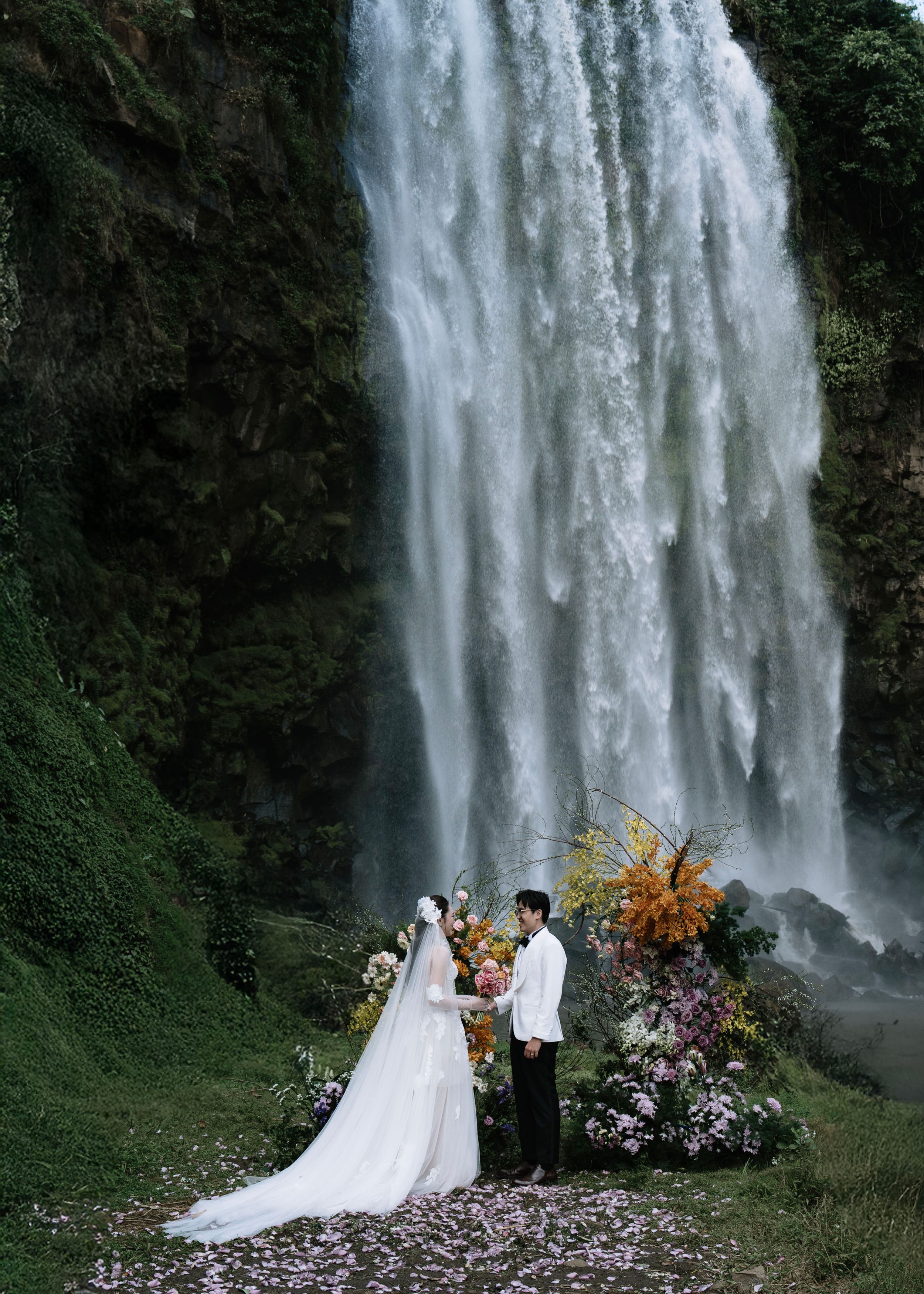 Bride and groom standing together before a dramatic waterfall surrounded by colorful floral arrangements.