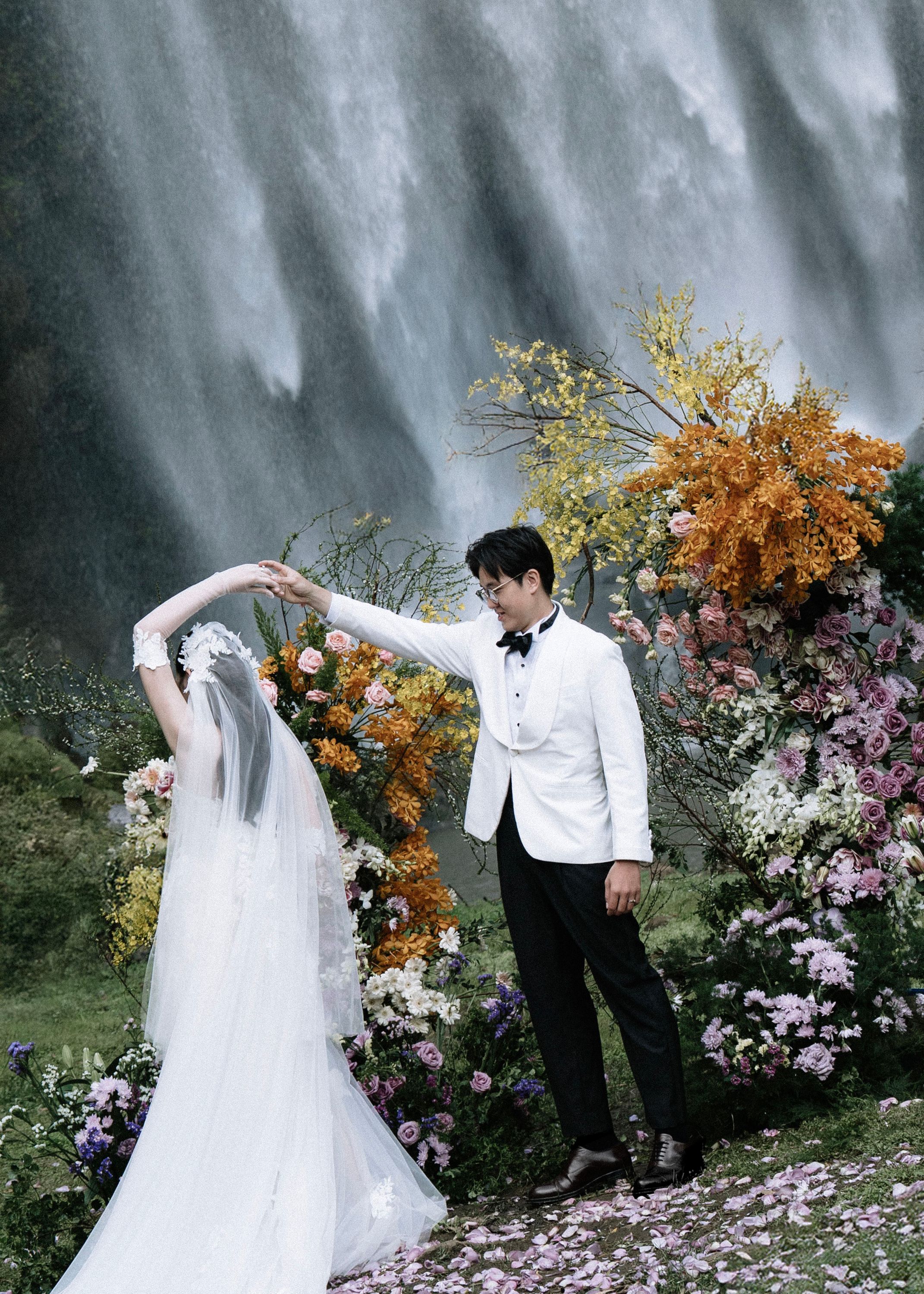 Groom spinning bride in elegant white gown before a waterfall surrounded by colorful blooms.