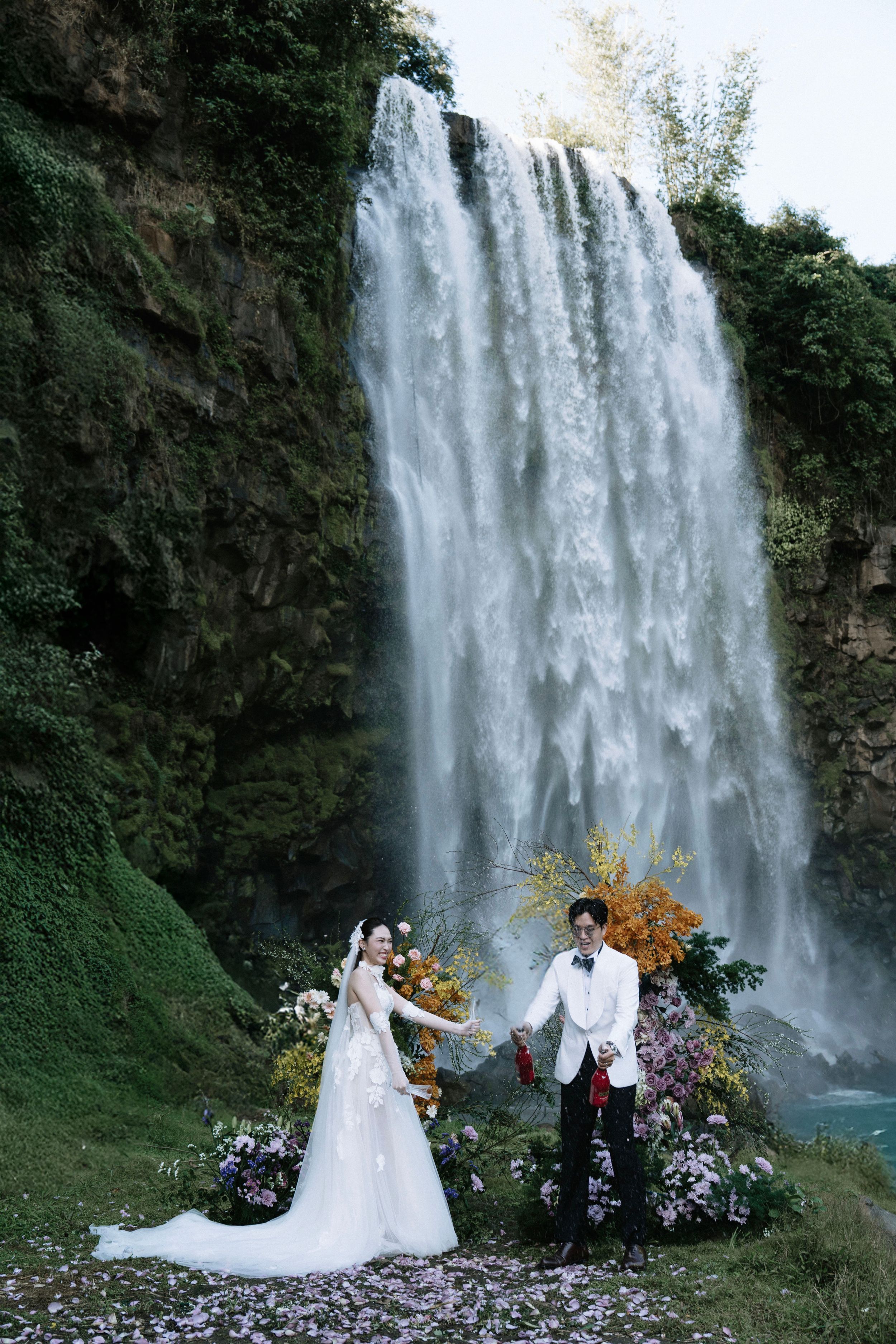 Couple exchanging vows before a majestic waterfall with vibrant floral arch and scattered petals.