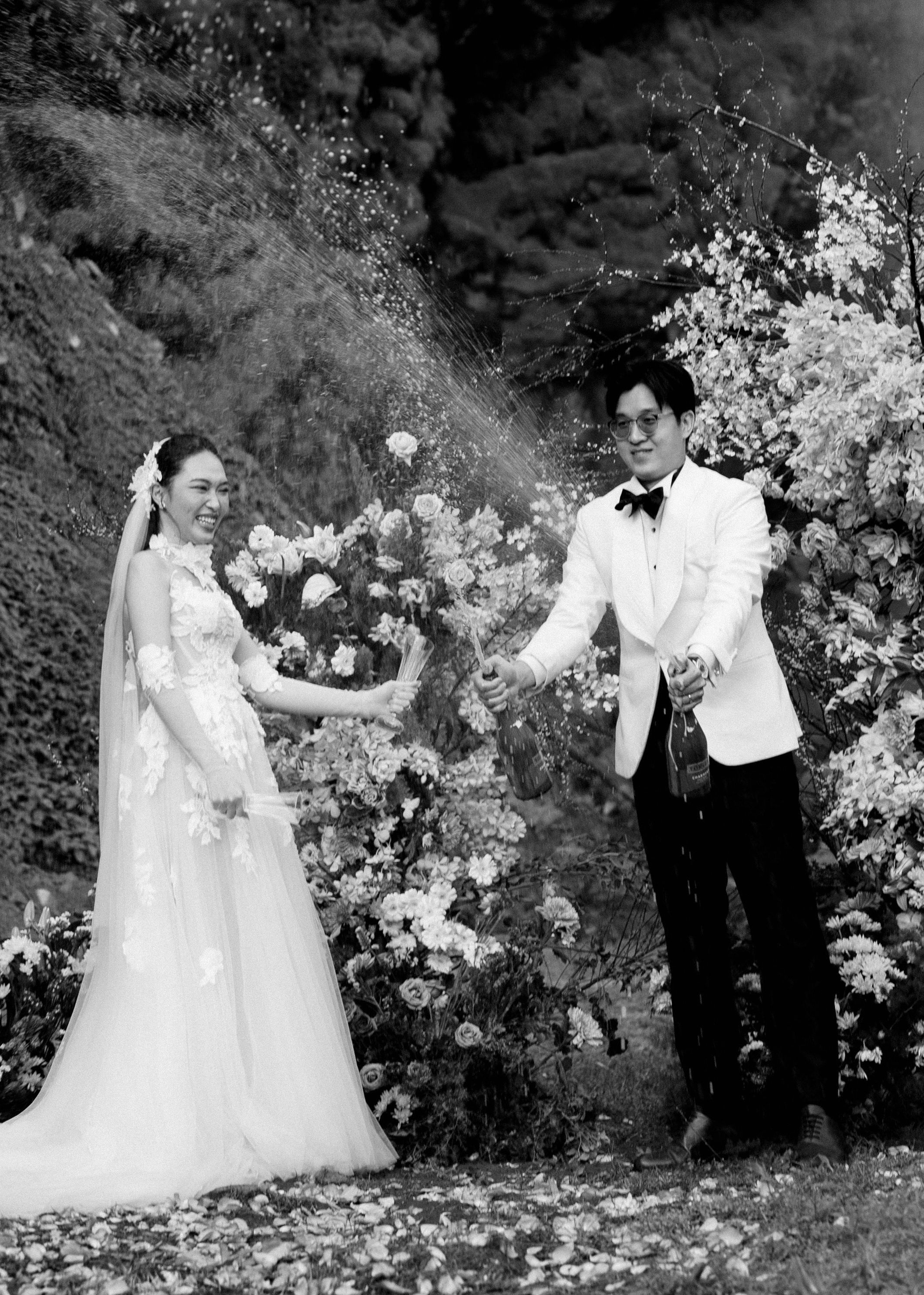 Black and white photo of laughing couple celebrating with champagne spray among garden flowers.