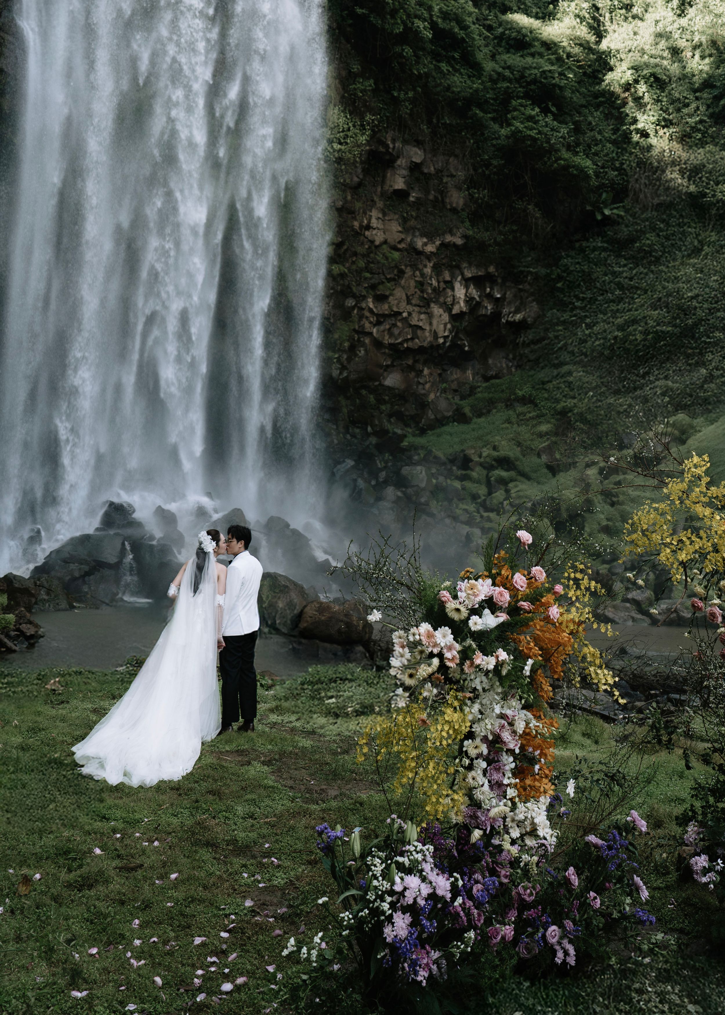 Bride and groom standing together before a majestic waterfall surrounded by colorful floral arrangements.