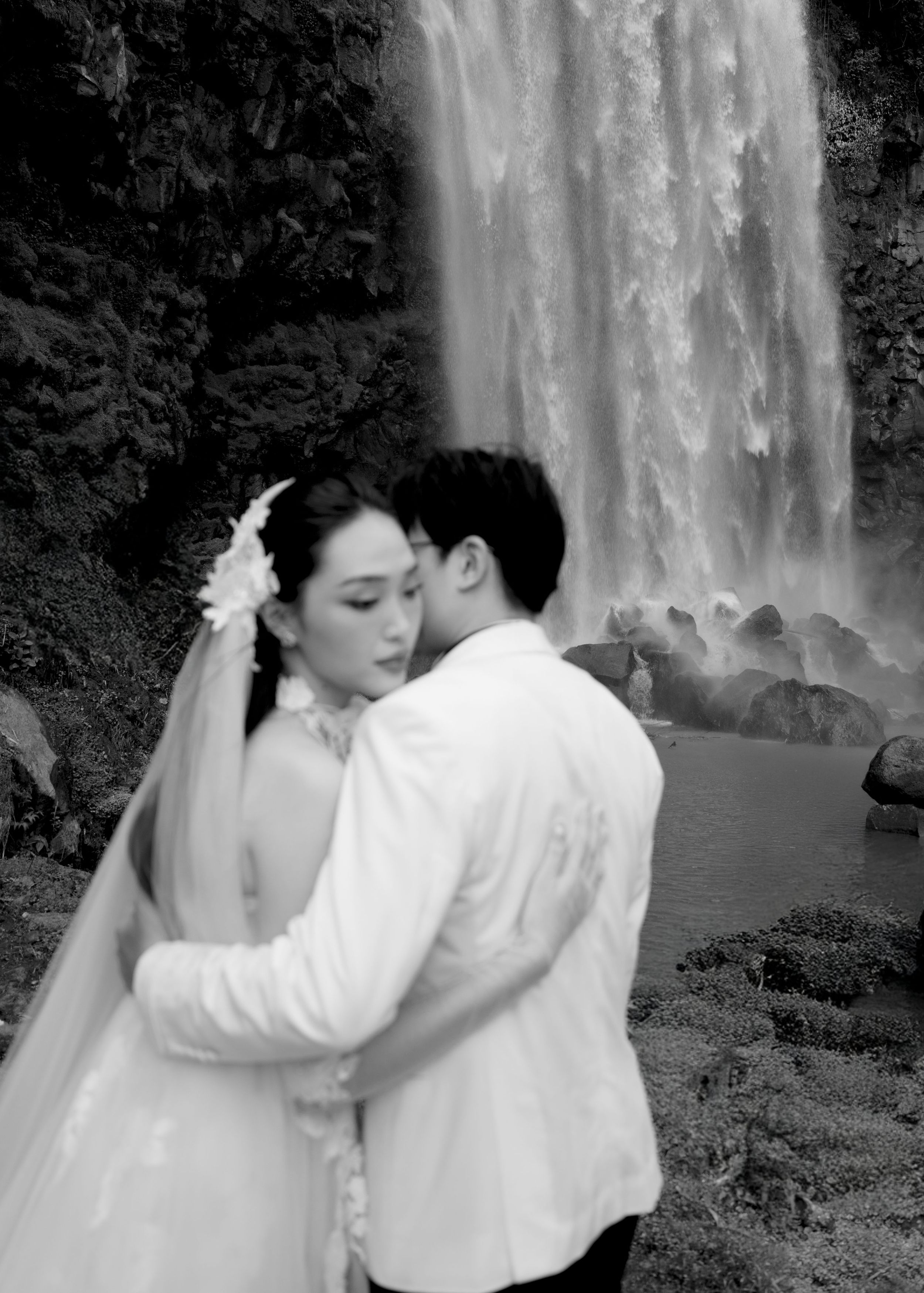 Black and white photo of bride and groom embracing romantically in front of a stunning waterfall.