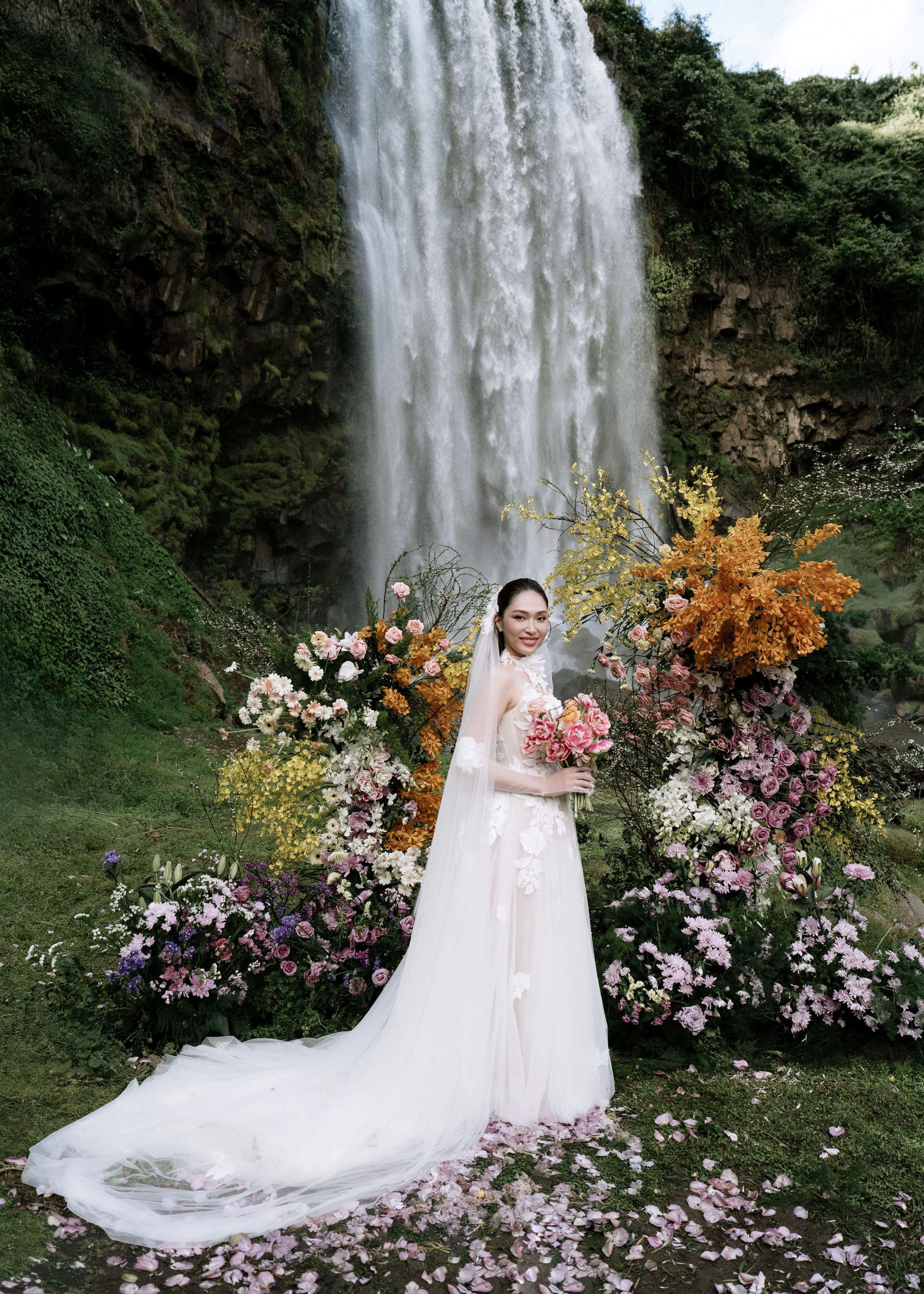 Elegant bride in white gown holding bouquet posing before waterfall with vibrant floral arch display.