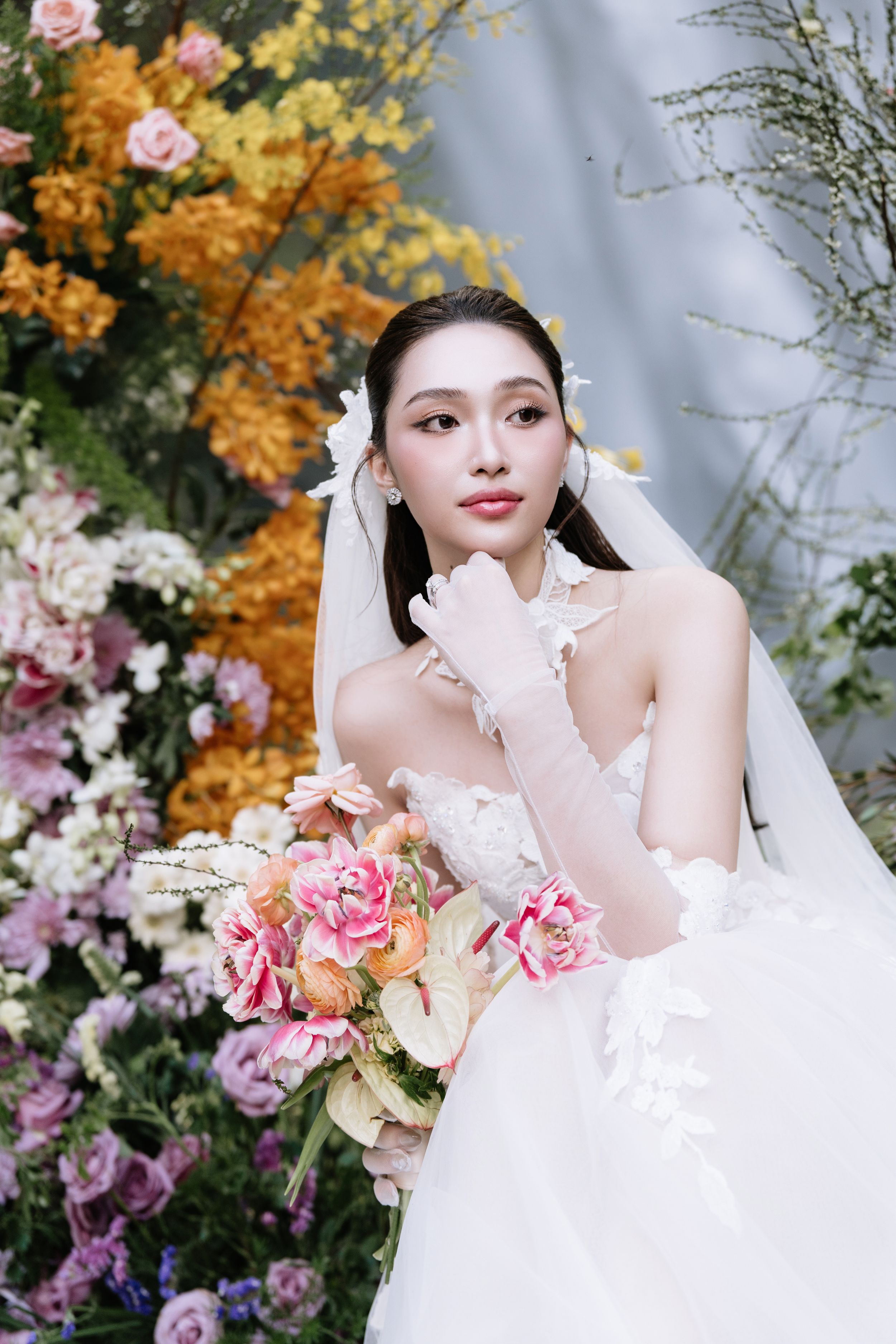 Elegant bride in white gown with veil and gloves posing among vibrant pink and yellow flowers.