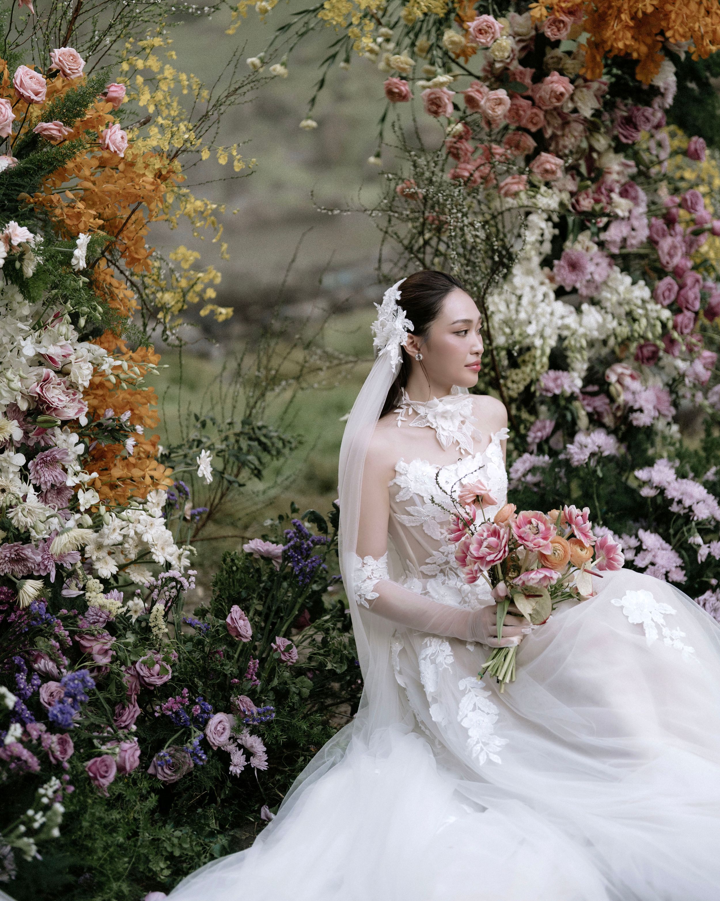 Beautiful bride in lace gown sitting among lush colorful garden flowers holding a pink bouquet.