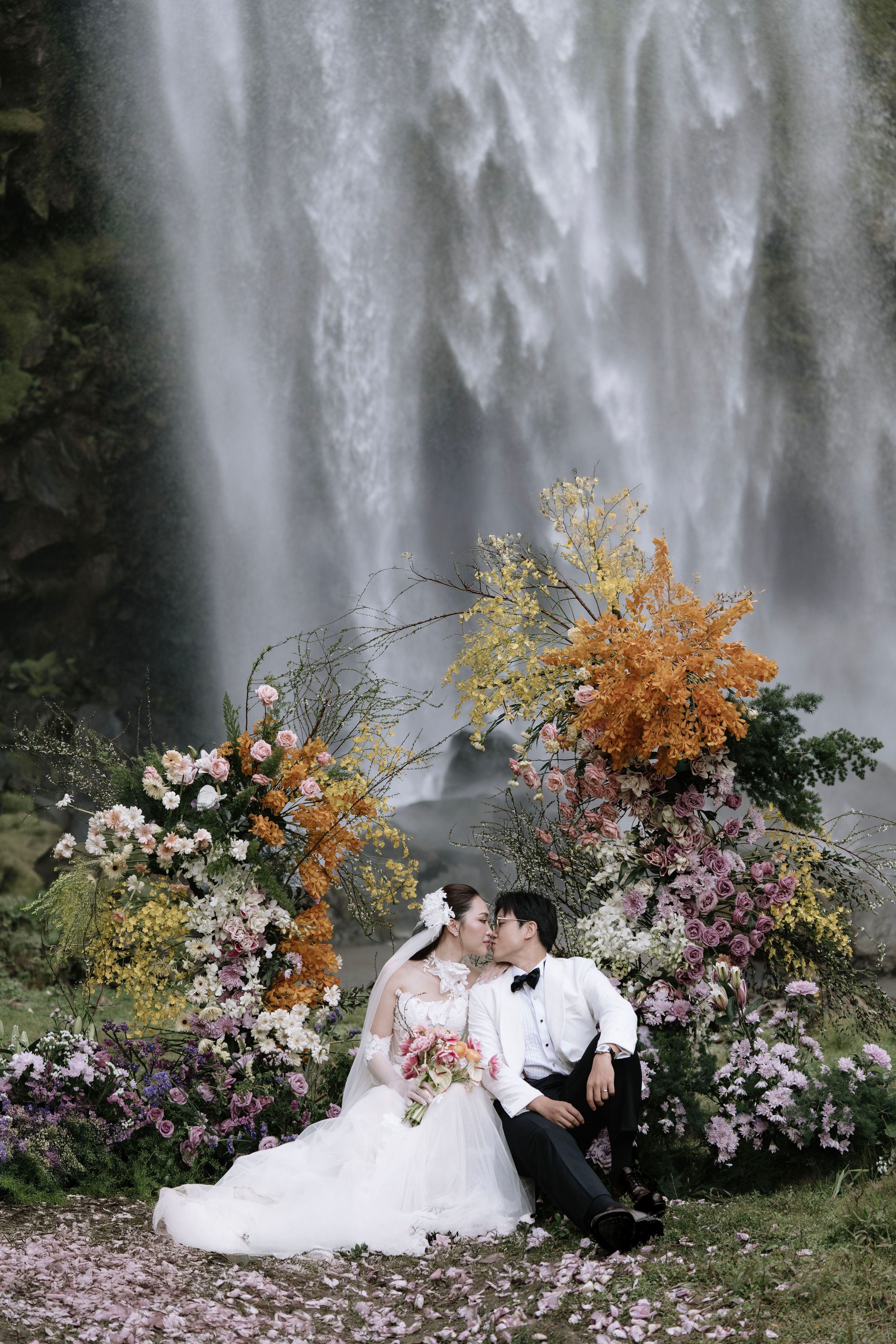 Bride and groom sharing a kiss seated before a waterfall surrounded by stunning floral arrangements.