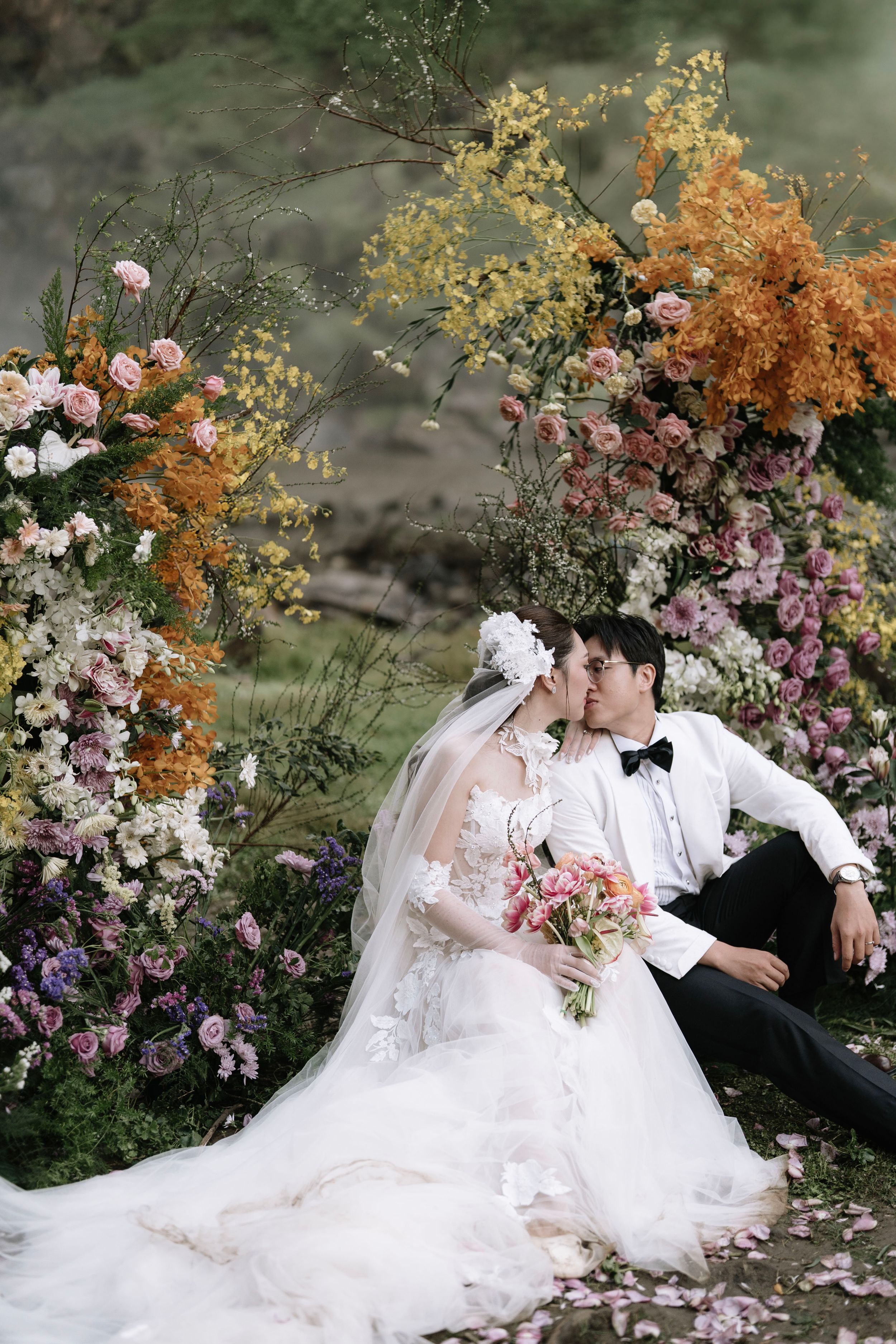 Bride and groom sitting together beneath a colorful floral arch with cascading waterfall background.