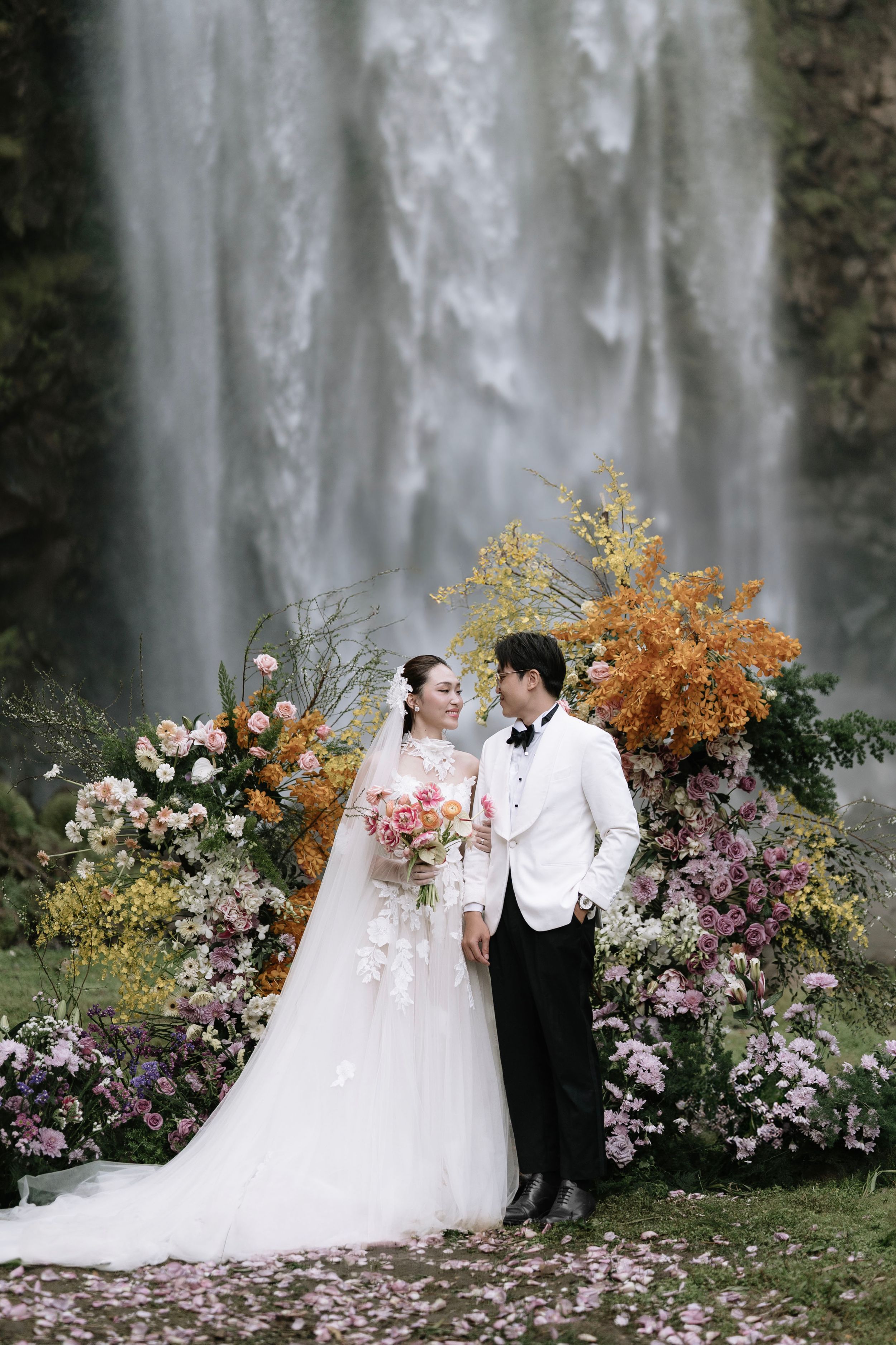 Bride and groom gazing at each other before a waterfall with colorful autumn floral backdrop.