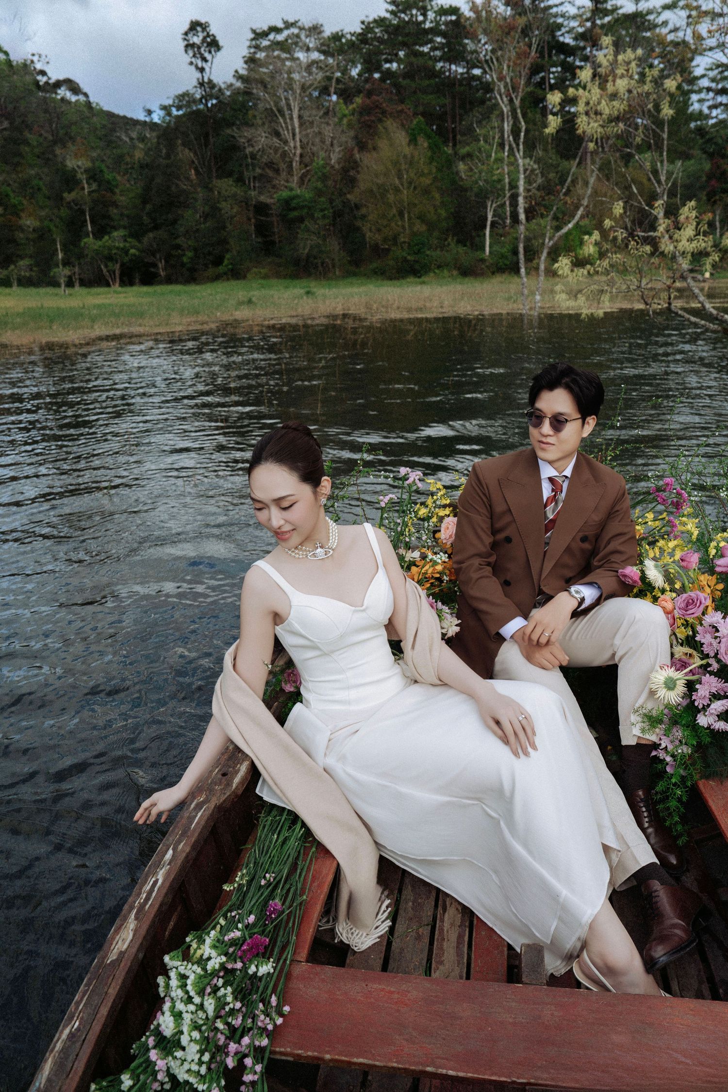 Bride in white slip dress and groom in brown suit sit in floral wooden boat on calm lake.
