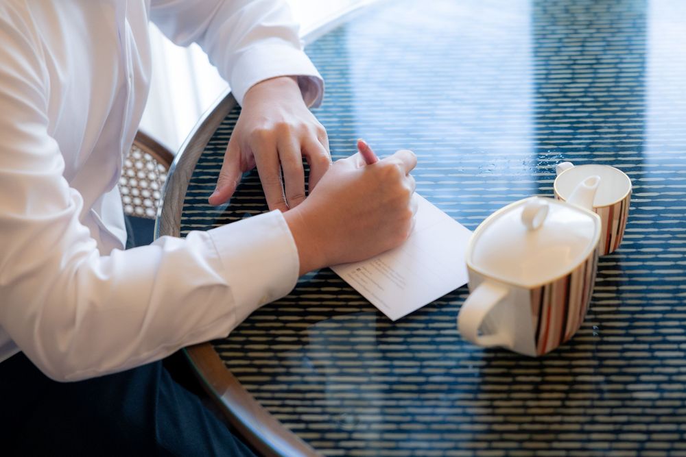 Someone wearing a white shirt writes notes in a notebook while sitting at a coffee table with a beverage.
