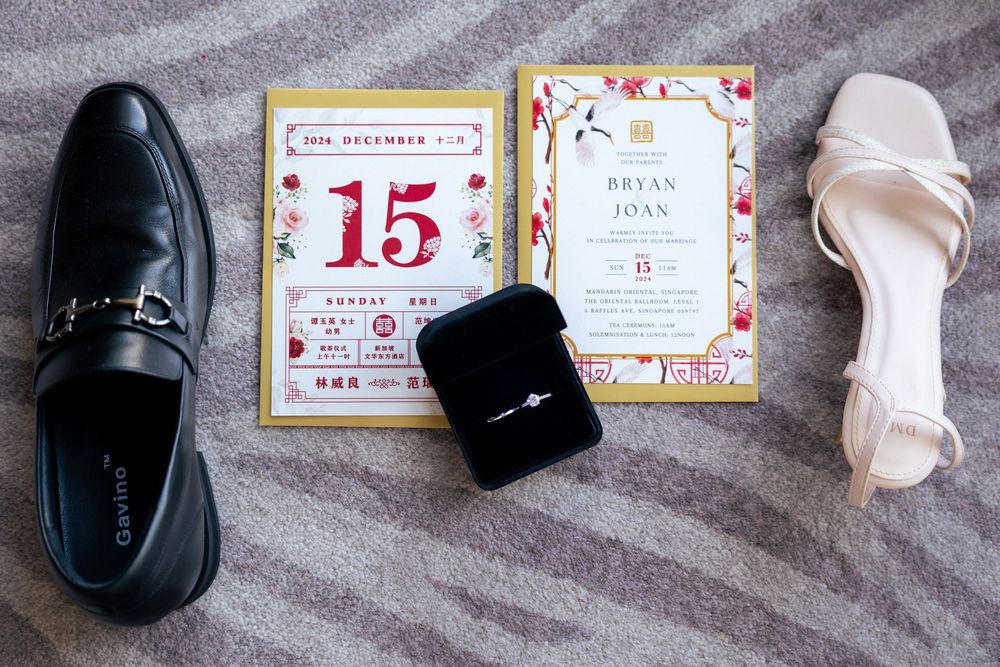 Wedding detail shot showing black dress shoe, invitation with number 15, ring box, and white bridal heel on gray carpet.