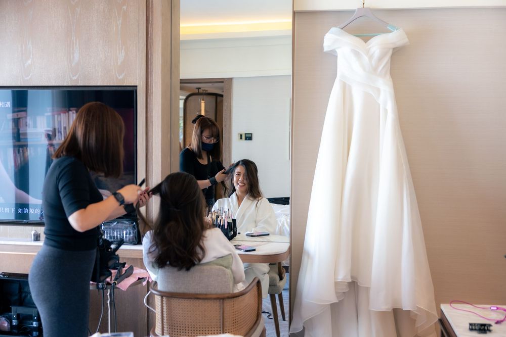 White wedding dress hangs in background while preparations take place in hotel room.