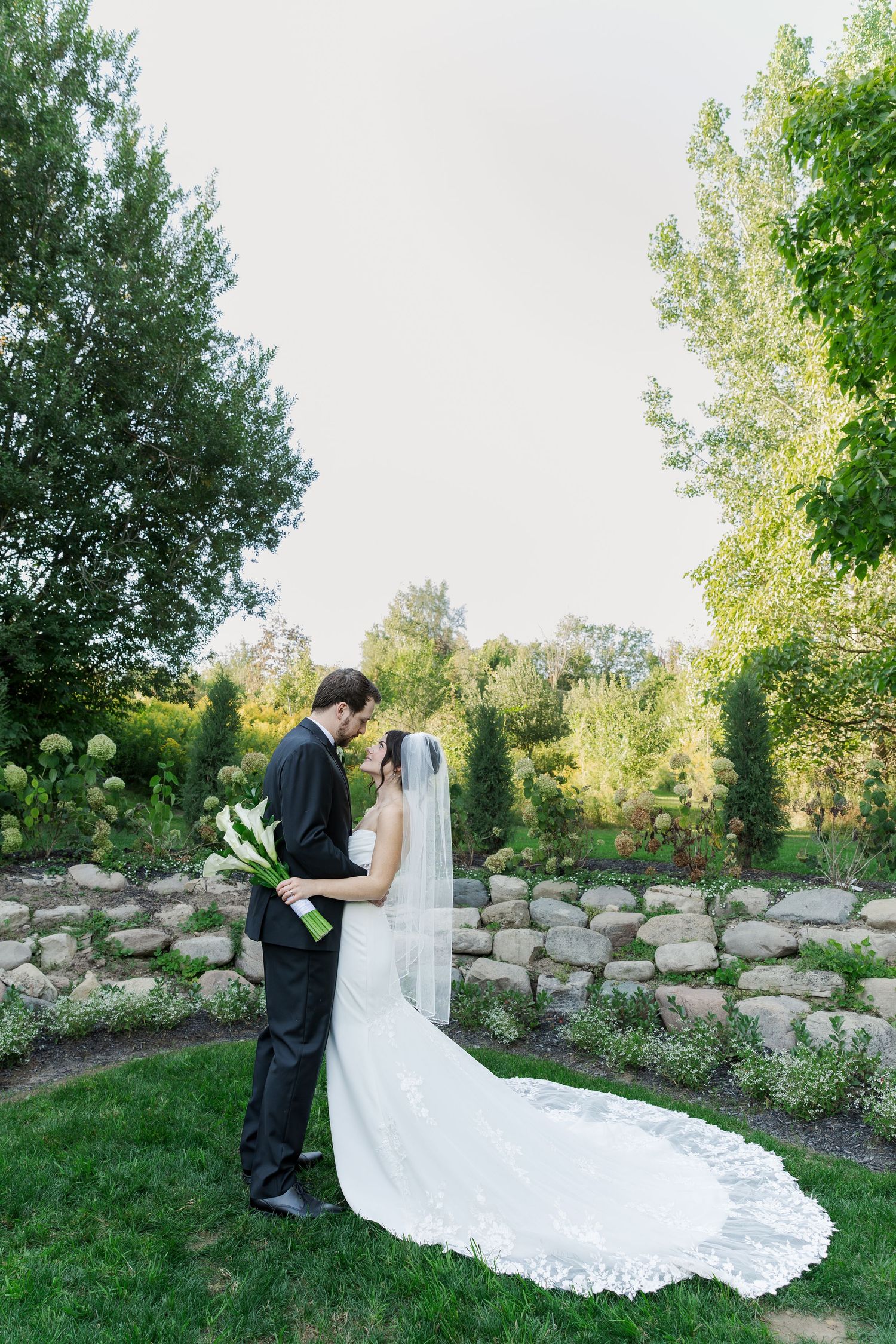 Bride's long lace train trails beautifully across green grass during outdoor wedding portraits at The Wren House in St. Jacobs, Ontario.