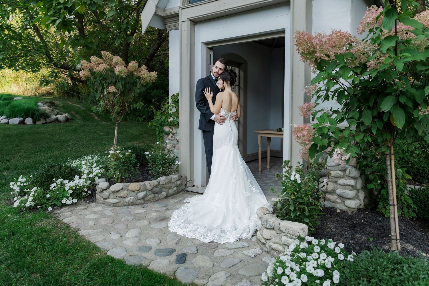 Bride and groom embrace in a white church doorway with stone foundation and flowering plants at The Wren House in St. Jacobs, Ontario.