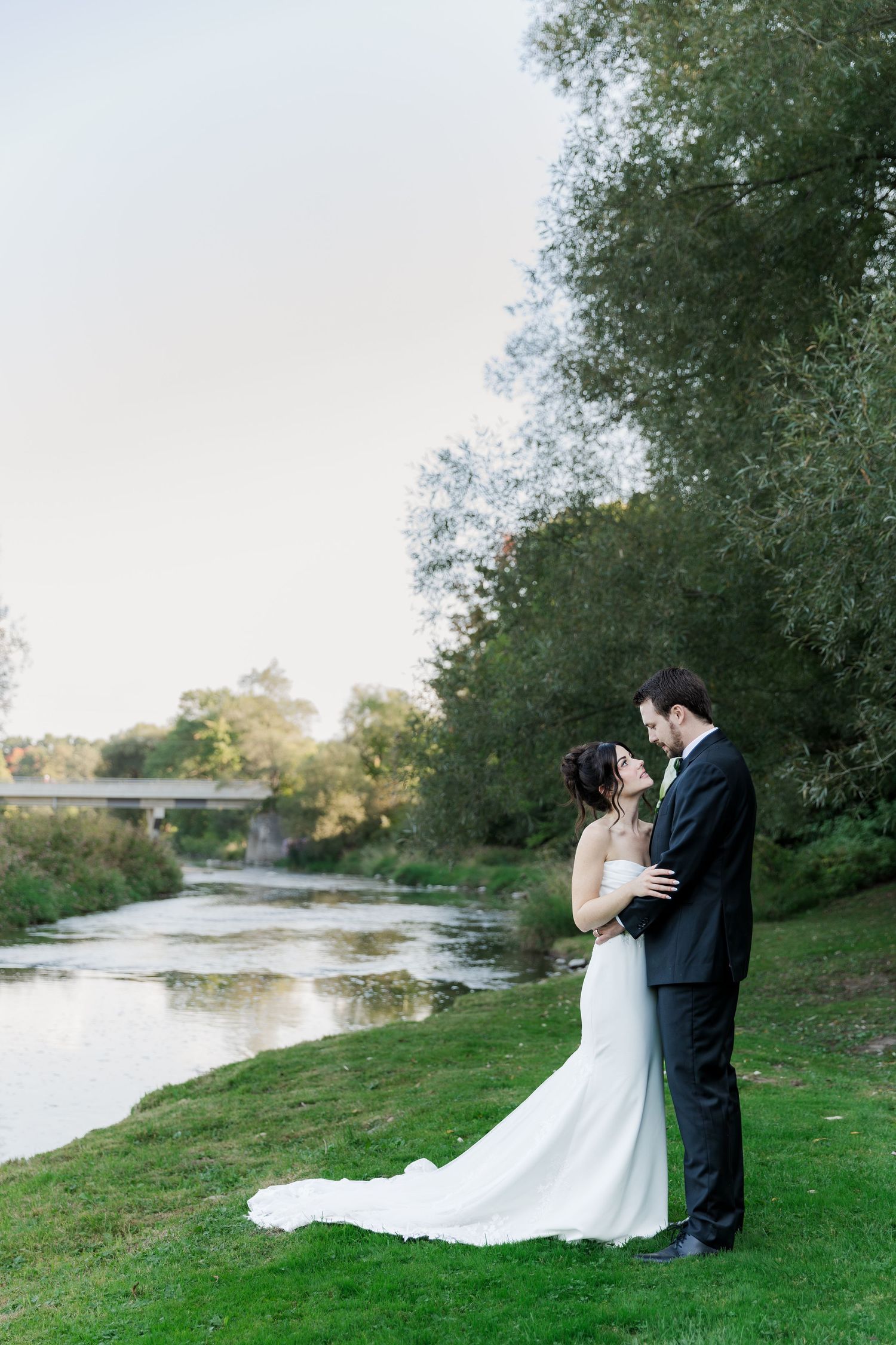 A couple shares an intimate moment by a river with trees and a bridge in the background at The Wren House in St. Jacobs, Ontario.
