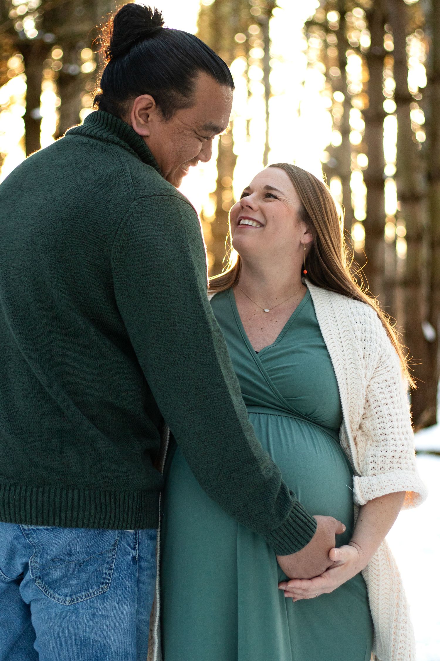 Maternity photo series showing couple in green sweaters and white cardigan against forest backdrop.
