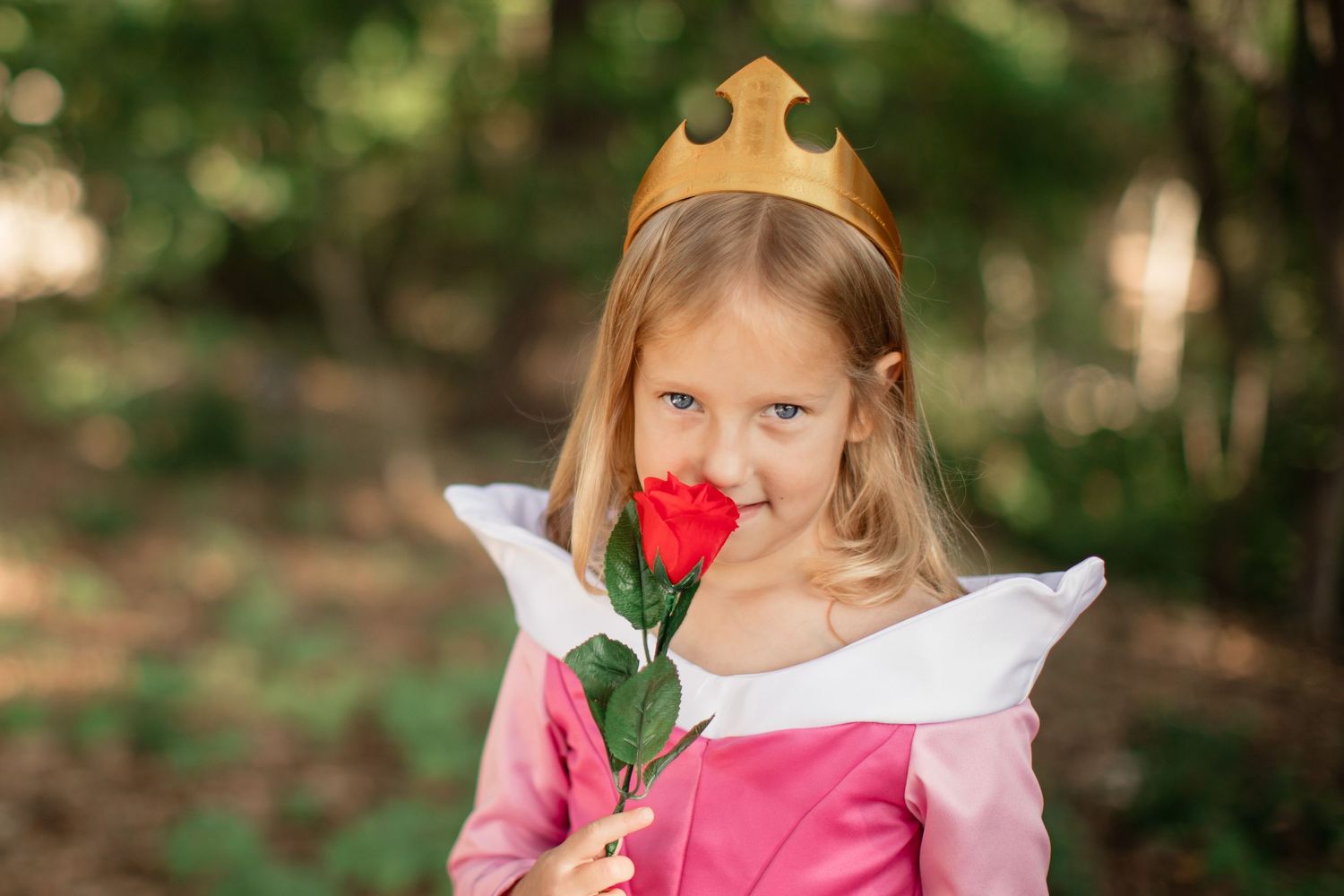 A young child in a pink princess dress and gold crown holds a red rose while standing in a forest setting.