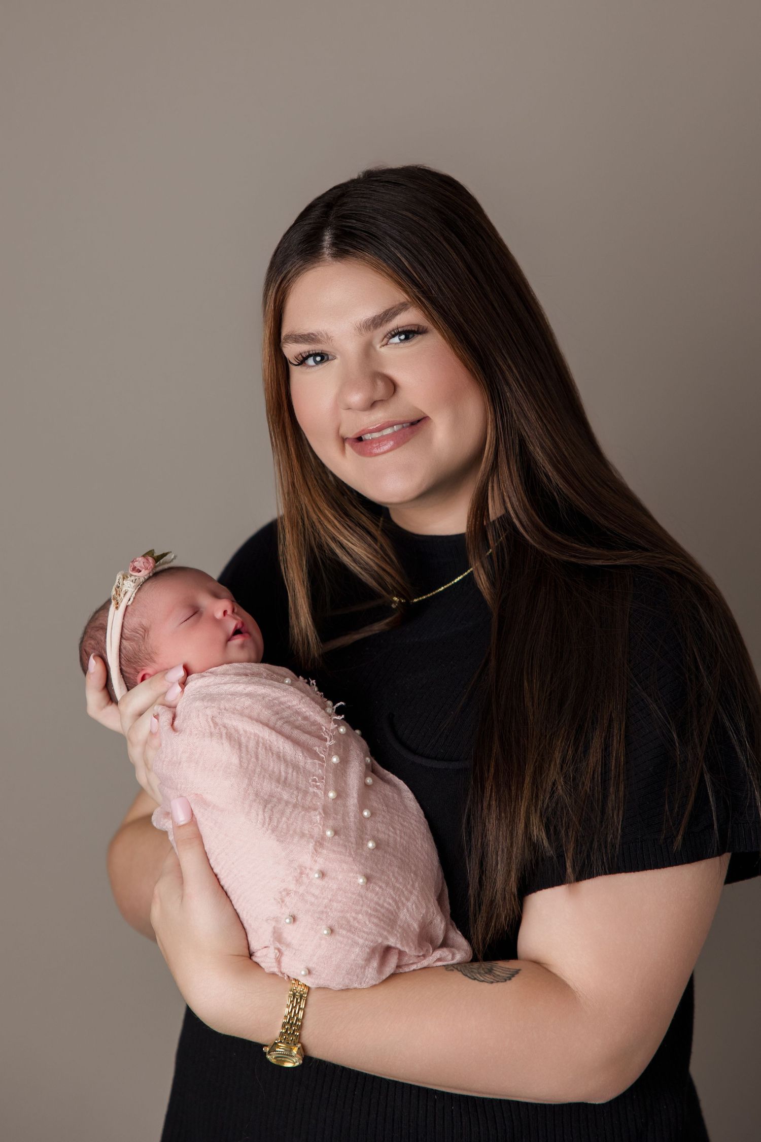 Woman in black shirt smiles while holding sleeping newborn baby wrapped in pink blanket for professional portraits.