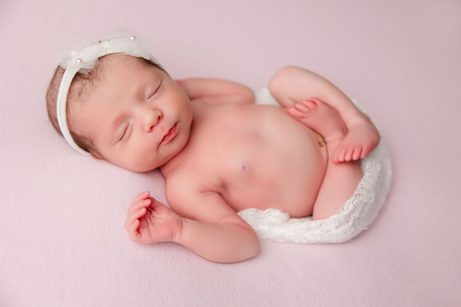 Sleeping newborn wearing white headband rests curled up on soft pink backdrop with delicate white wrap.