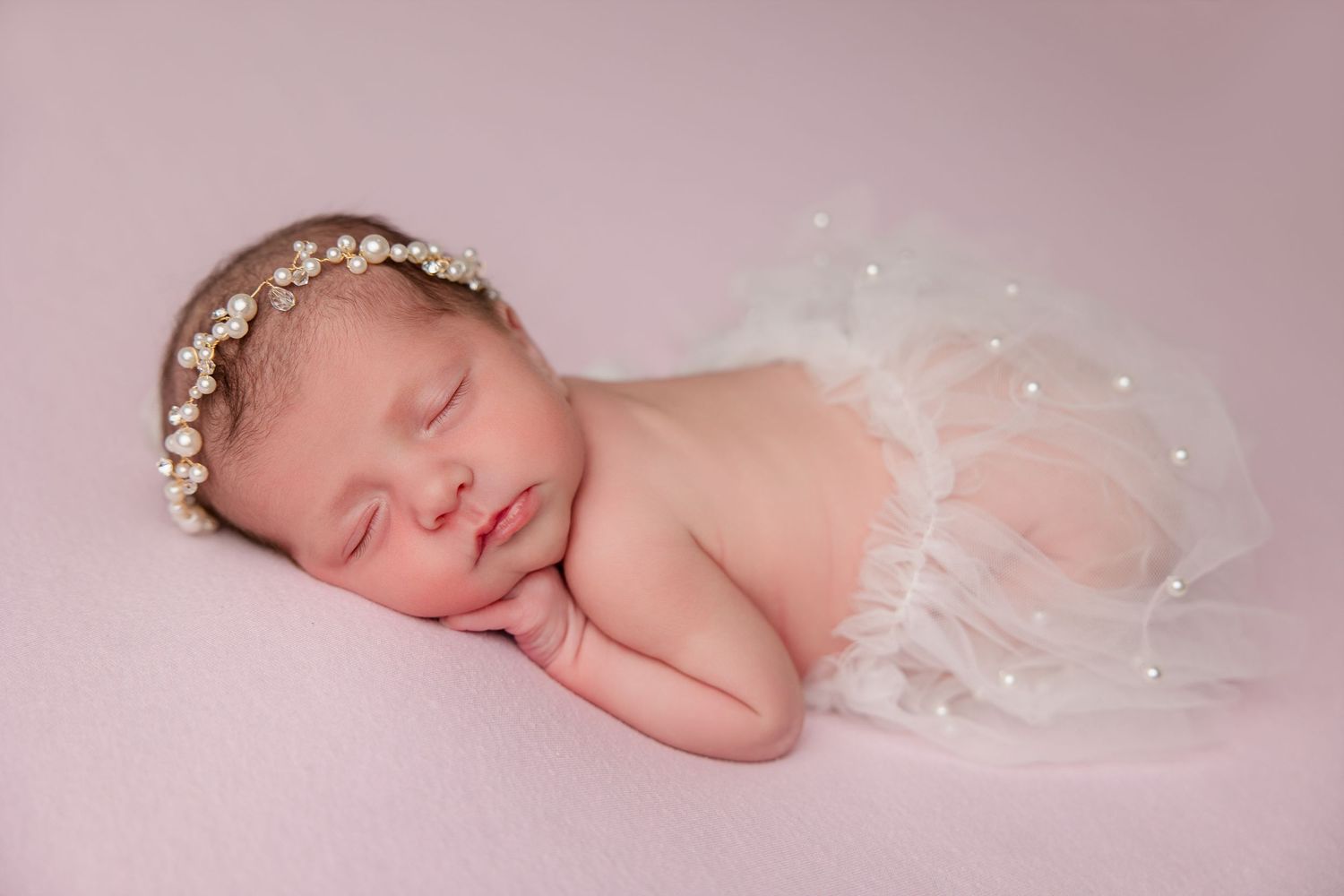 Sleeping newborn wearing delicate floral crown and tulle wrap on pink backdrop.