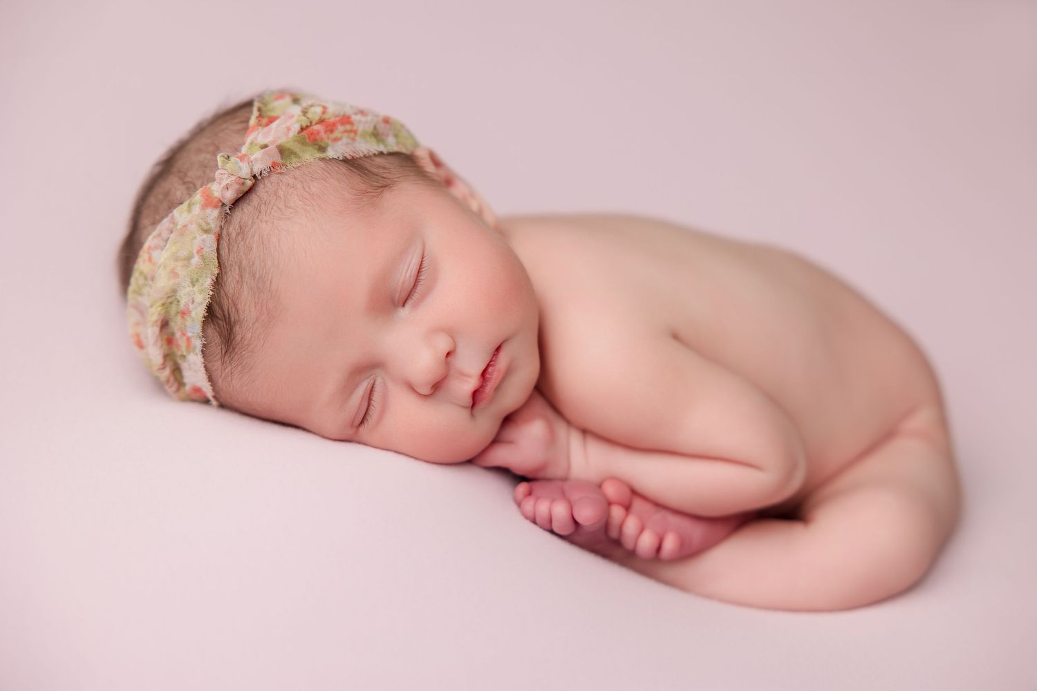 Peaceful newborn sleeps on pink background wearing floral headband in curled pose.