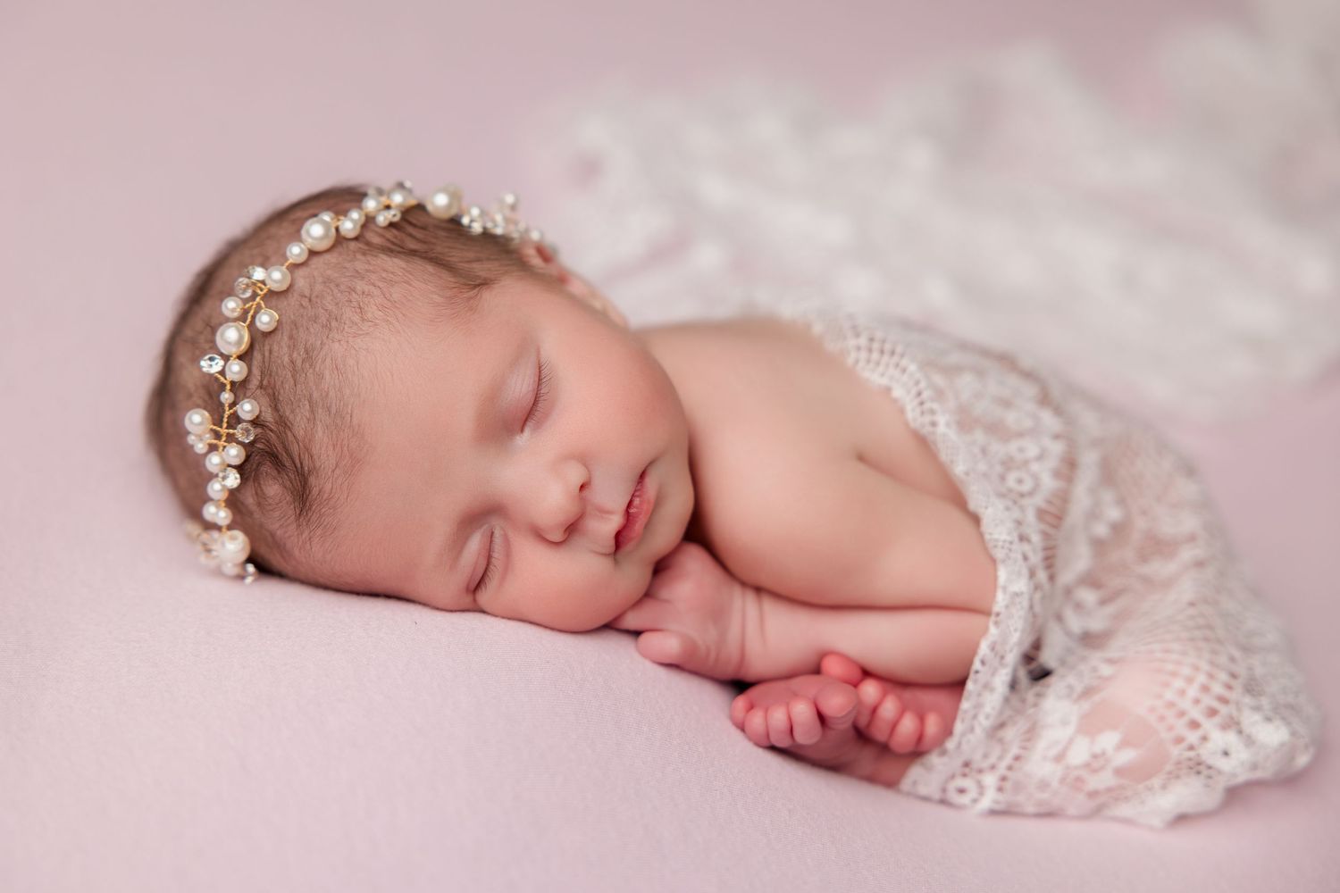 Dreaming newborn with pearl crown rests on pink backdrop draped in delicate lace.
