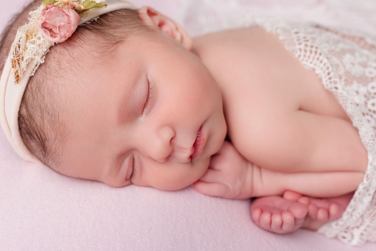 Sleeping newborn baby wrapped in delicate lace on pink background during photo session.