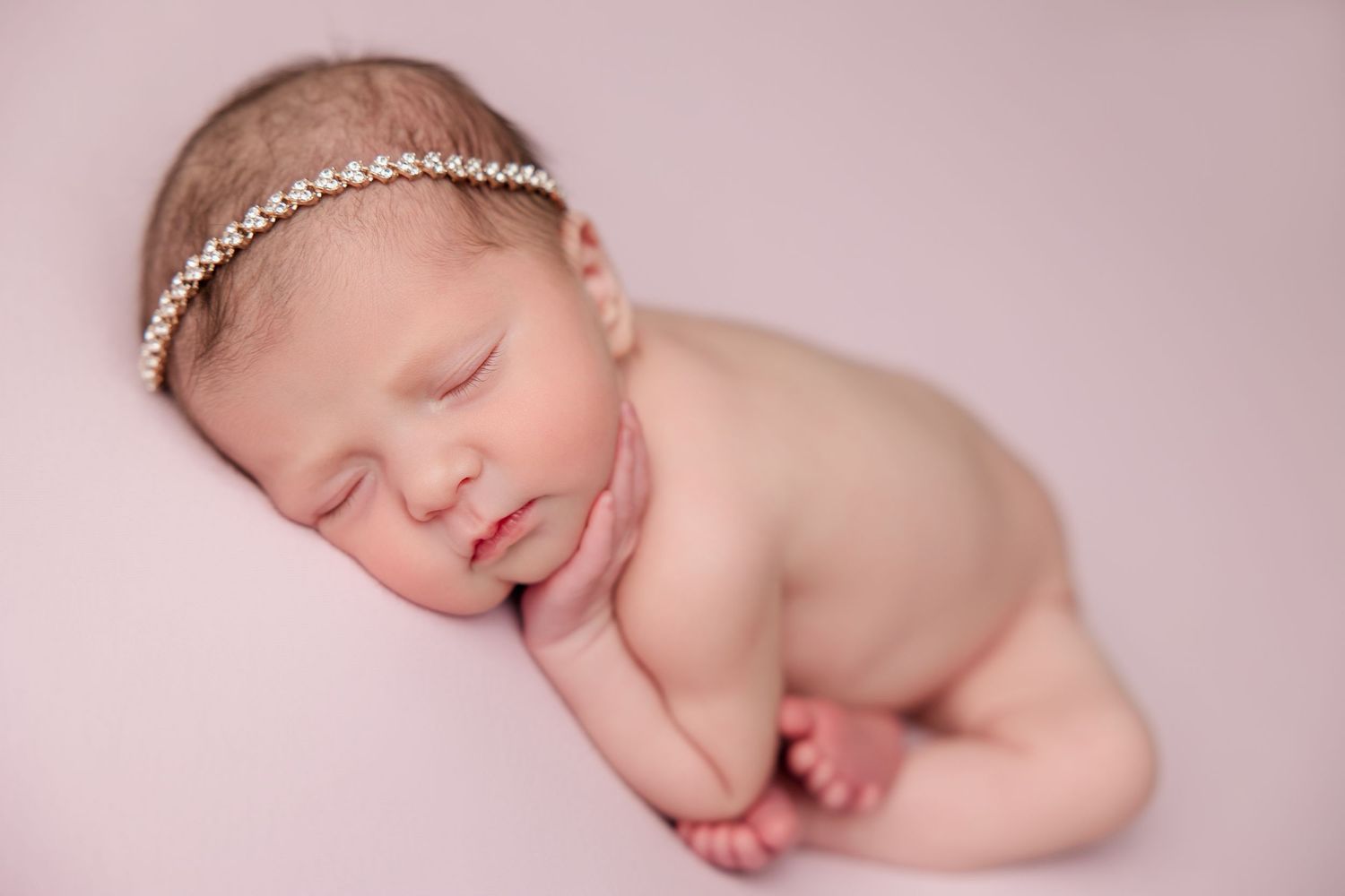 Sleeping newborn in delicate pink tones wearing a pearl headband during professional studio photography session.