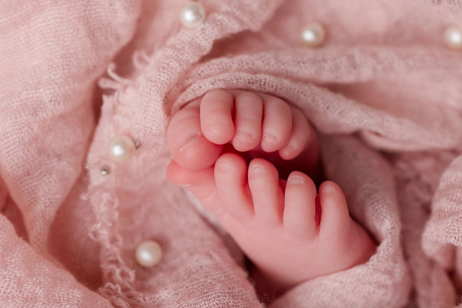 Close-up of tiny baby feet resting on pink fabric decorated with pearl embellishments.