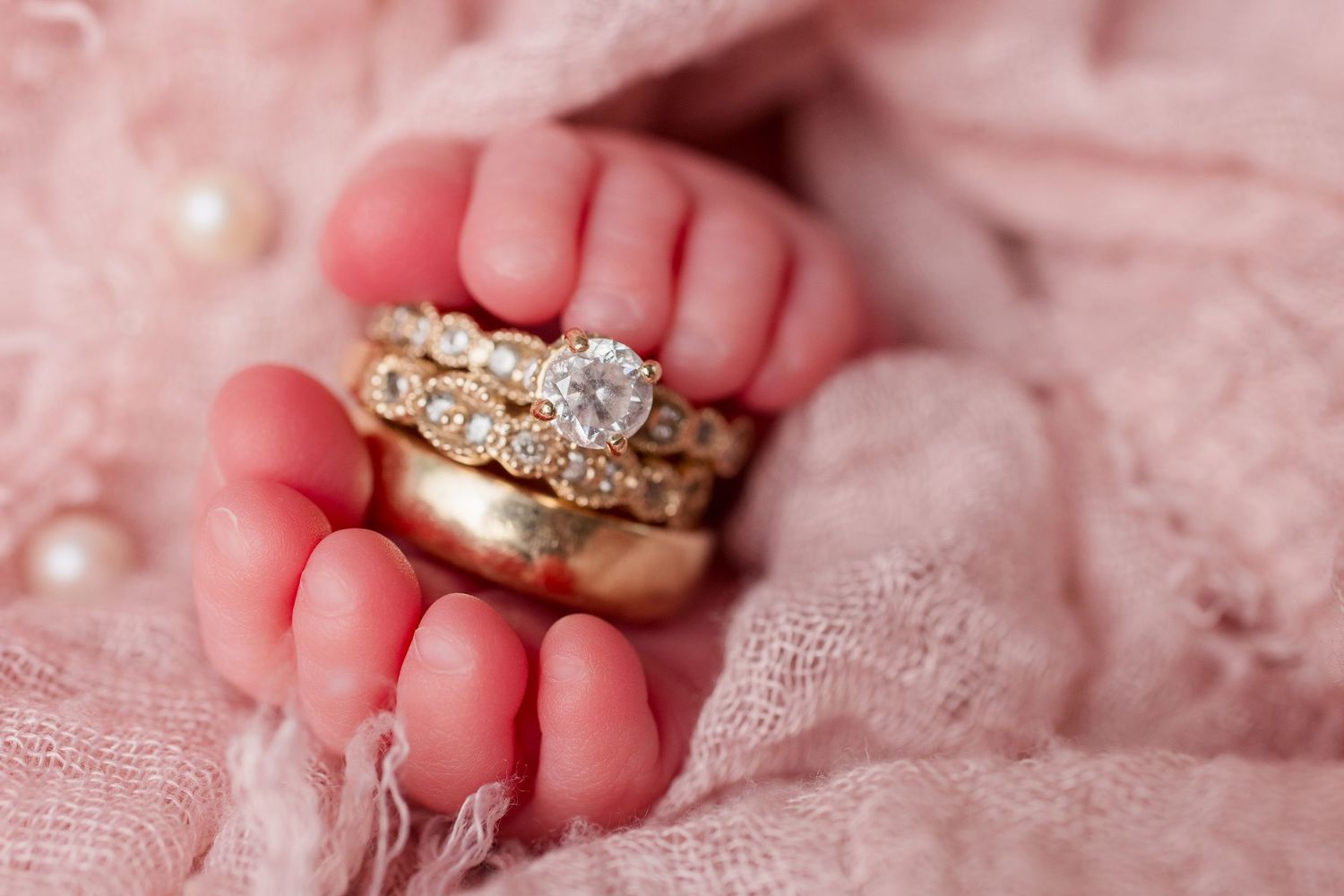 Close-up of tiny baby fingers holding an ornate pearl and diamond ring on pink textured fabric.