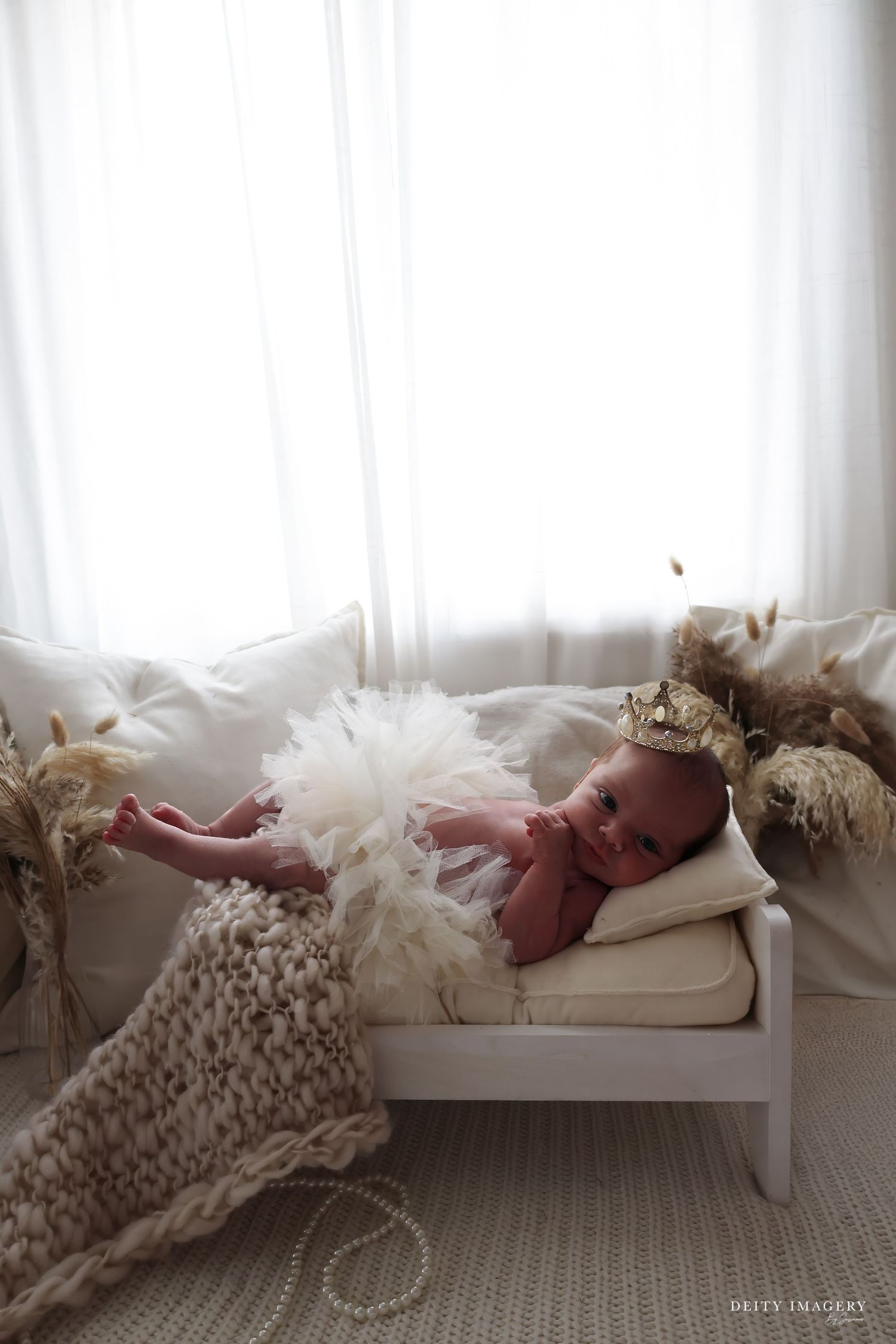 Newborn baby wearing a gold crown and white feathers lies on a tiny white bed with soft blankets near a bright window.