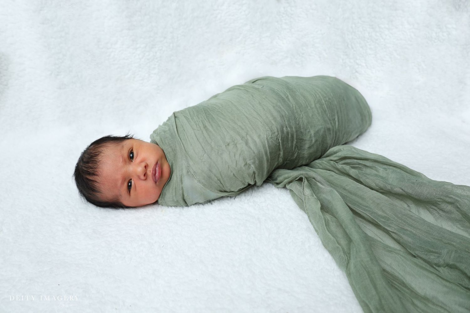 Newborn baby swaddled in a soft sage green wrap, lying peacefully on a white fluffy blanket.
