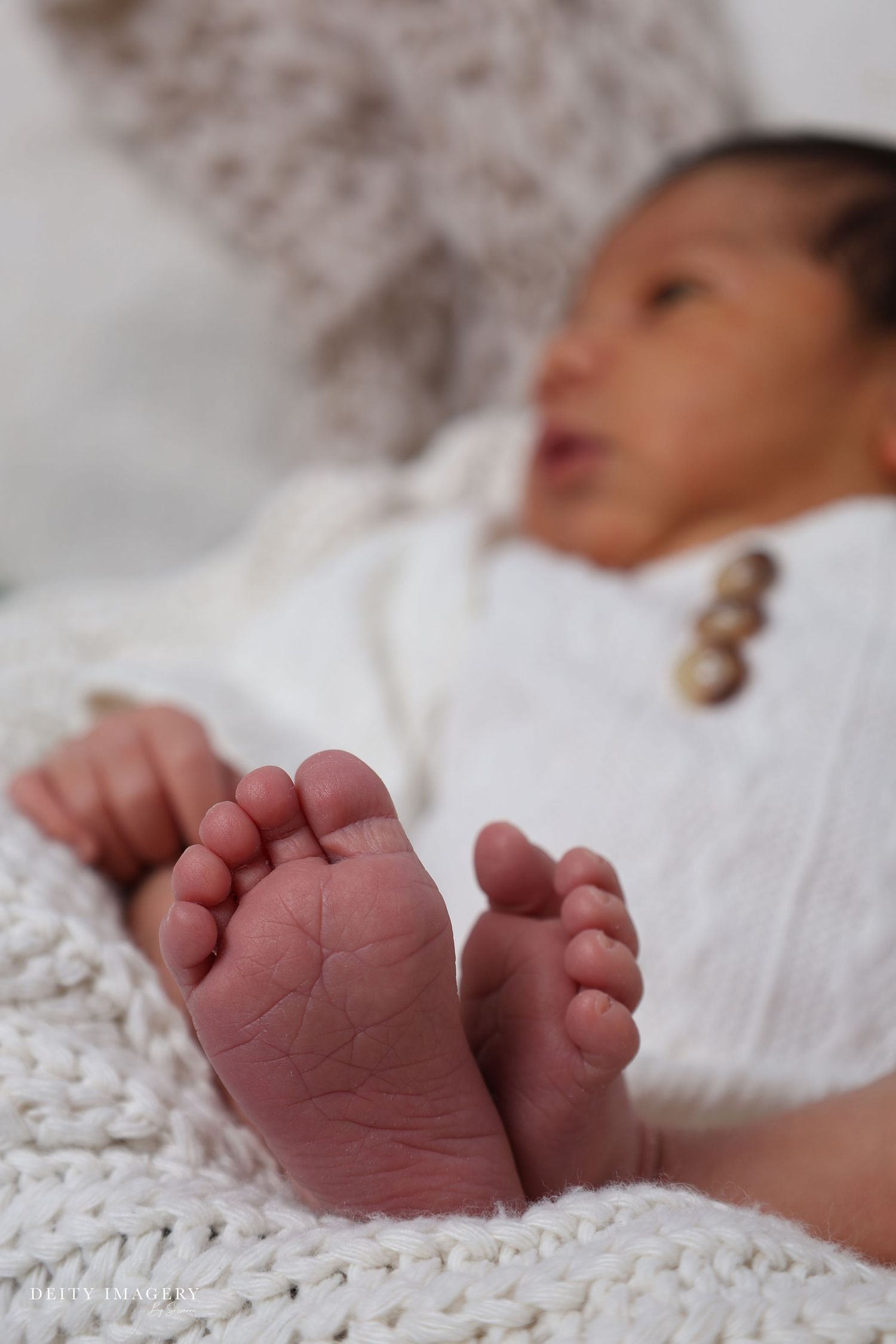 Close-up of tiny newborn baby feet on a white knit blanket with blurred infant in background.