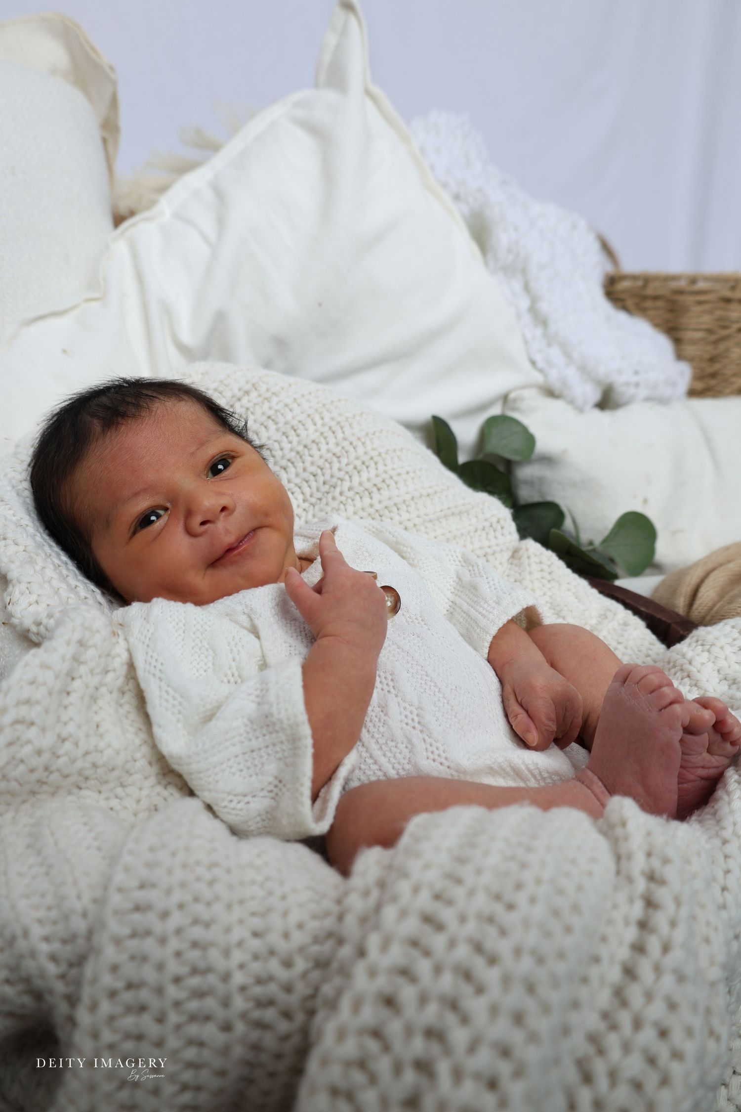 Newborn baby in white knit outfit resting on soft white blankets, smiling softly during a professional newborn photo session.