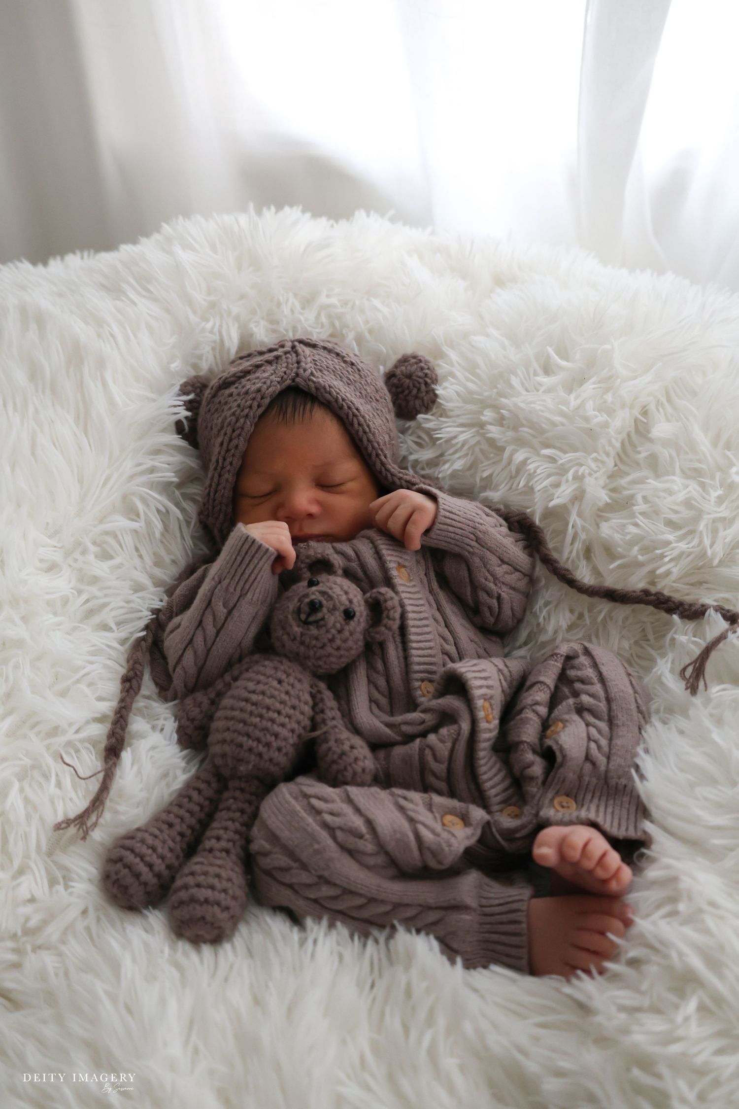 Sleeping newborn baby in brown knit outfit and hat cuddles a crocheted teddy bear on a white fluffy blanket.