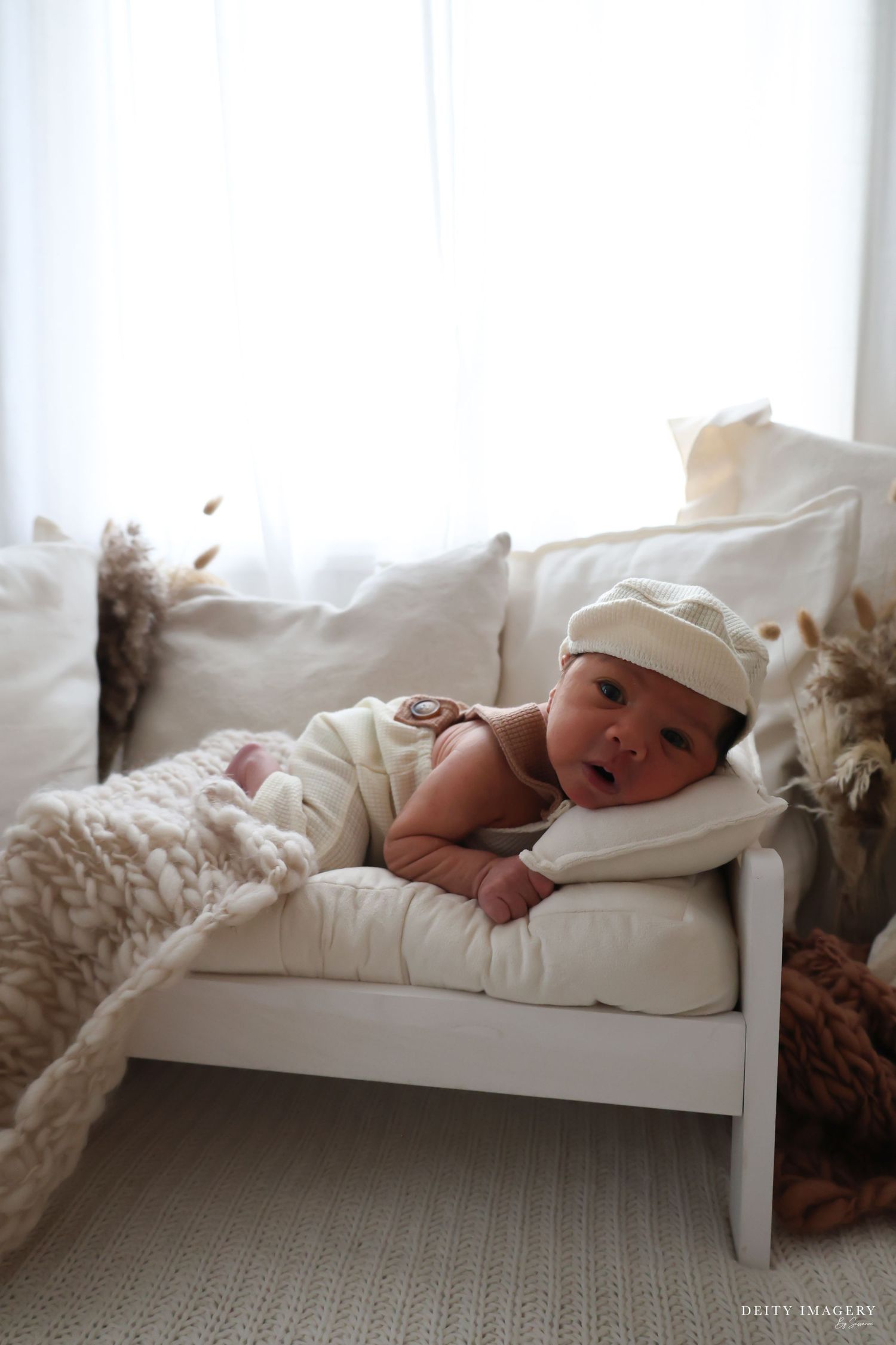 Newborn baby in a white cap resting on a tiny white couch with cream pillows and a chunky knit blanket.
