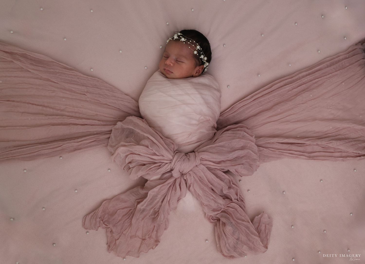 A newborn baby wrapped in white swaddle lies on pink tulle fabric arranged in a butterfly wing pattern.