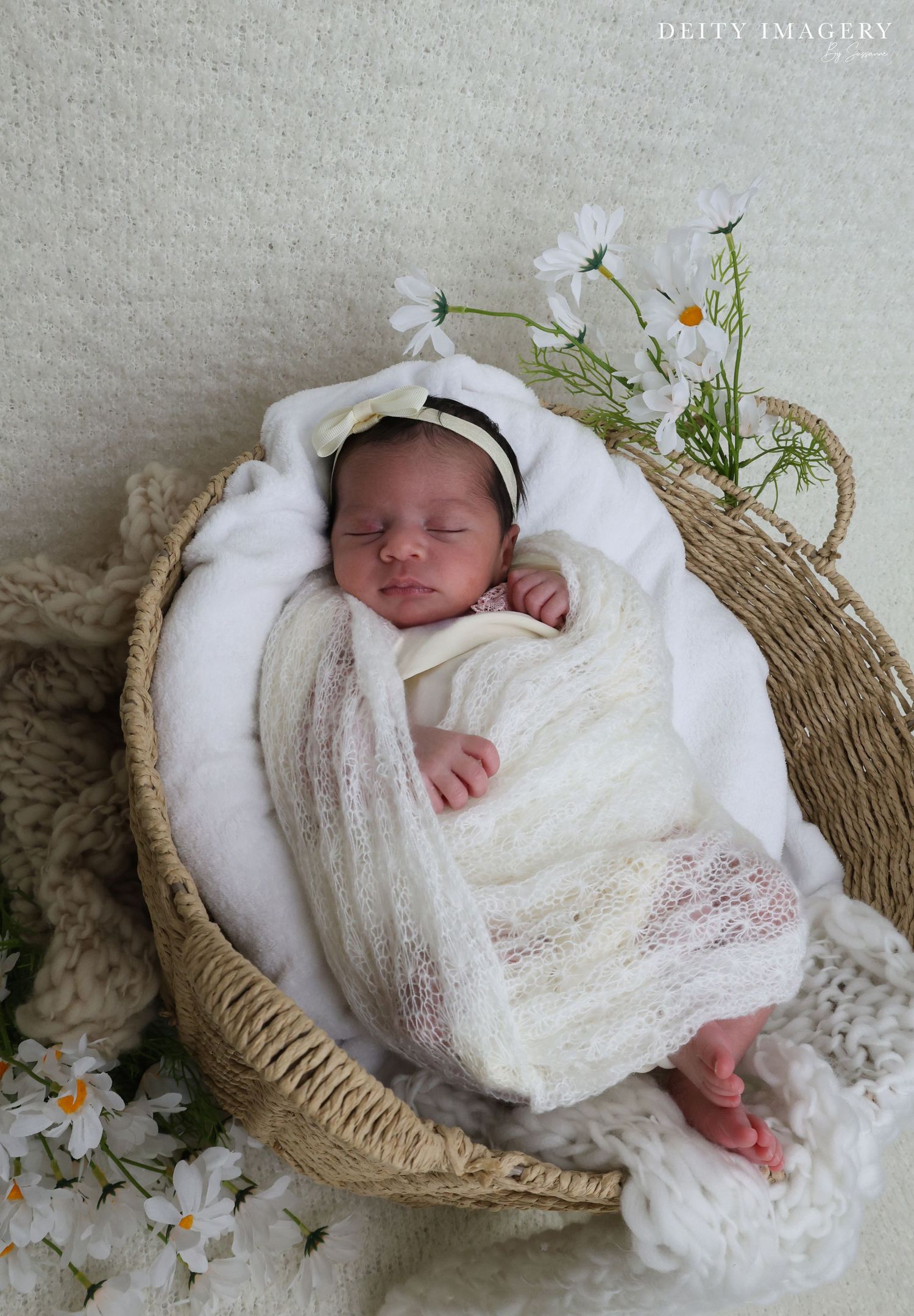 A sleeping newborn wrapped in white lace swaddle rests peacefully in a woven basket with delicate daisies as decoration.