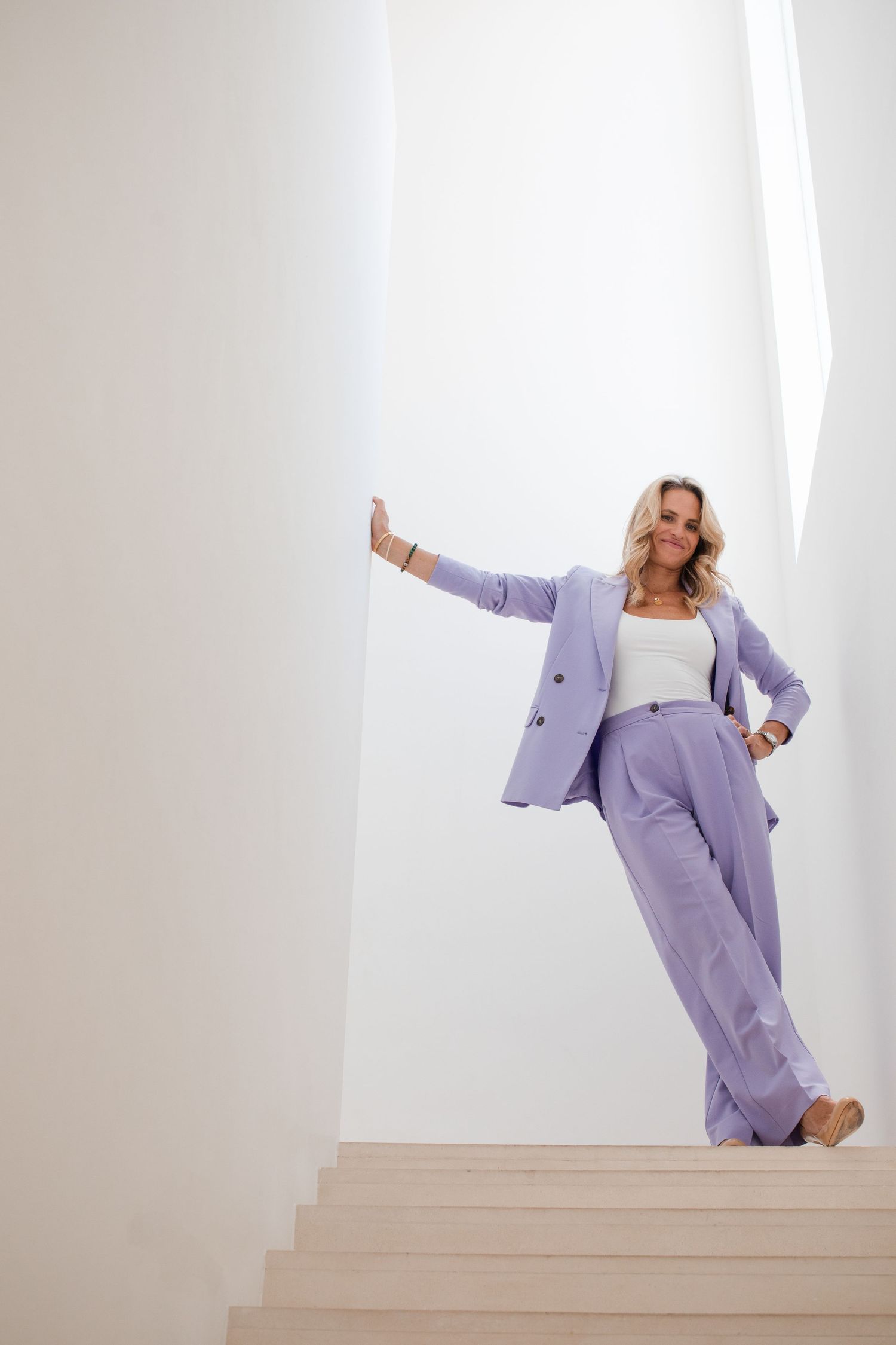 Woman in lavender pantsuit poses elegantly on white stairs in a bright minimalist space.