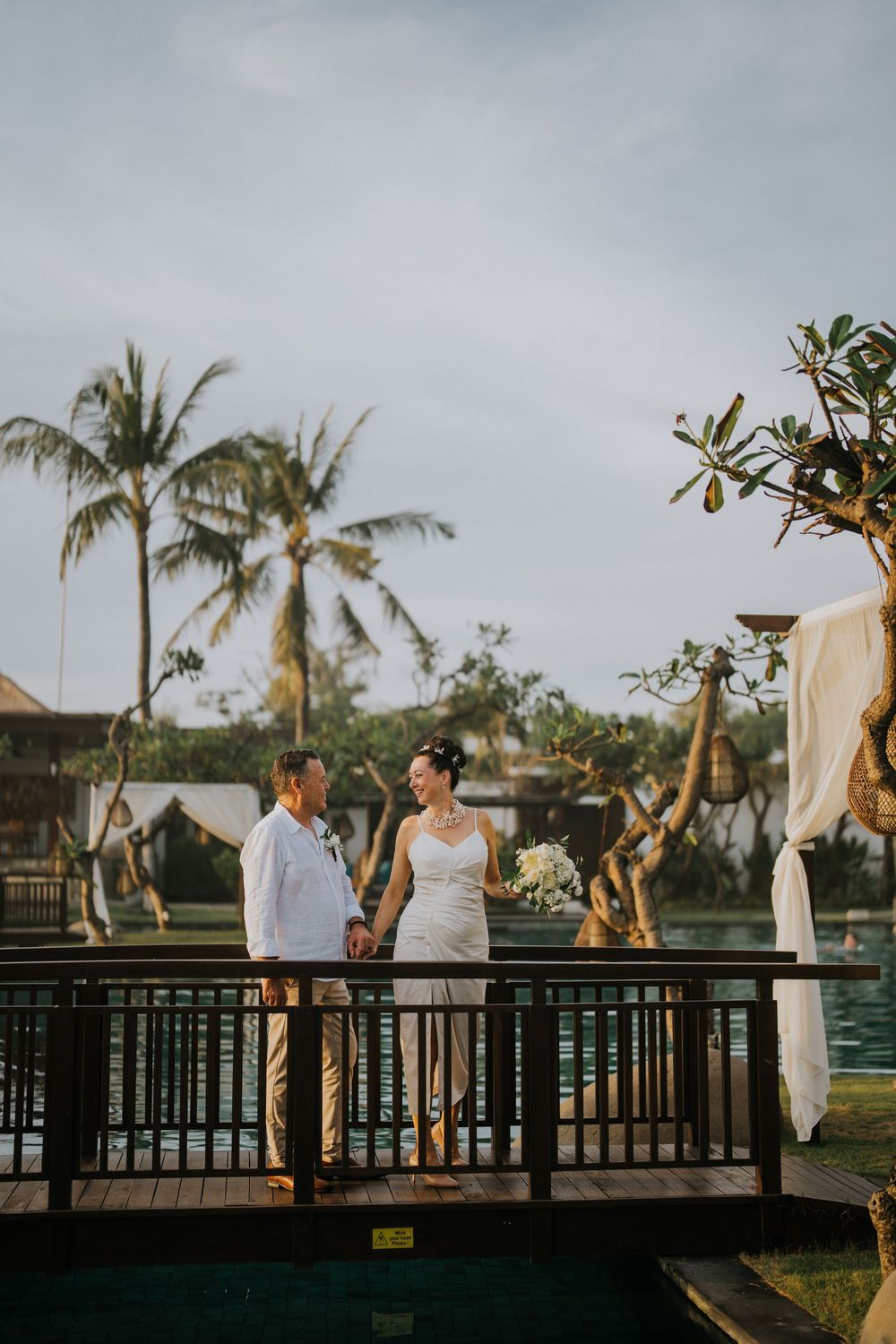 Wedding reception setup on a wooden deck with palm trees and ocean views at dusk.