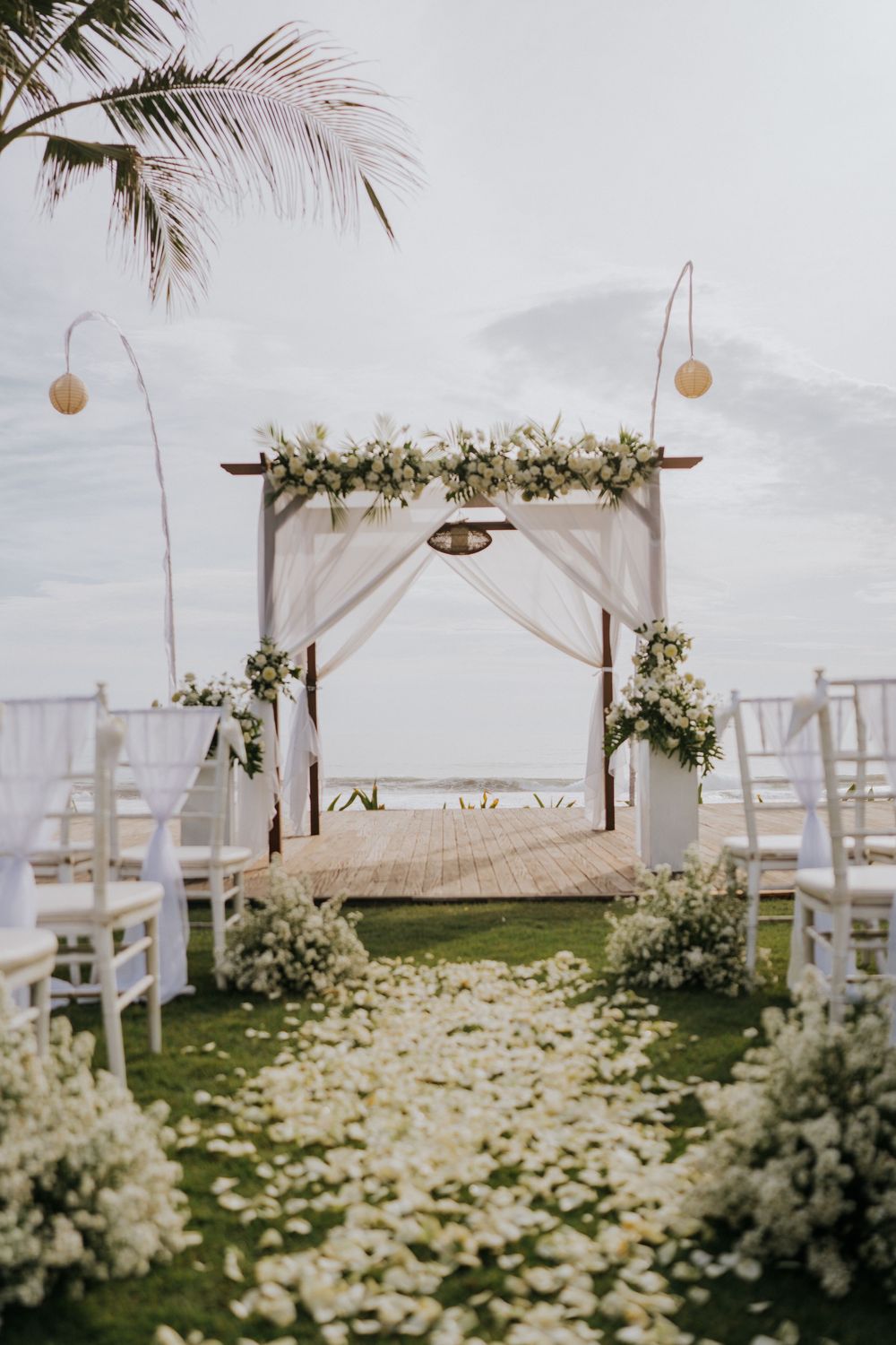 Beautiful floral arch and white chairs set up for beach wedding ceremony with scattered petals.