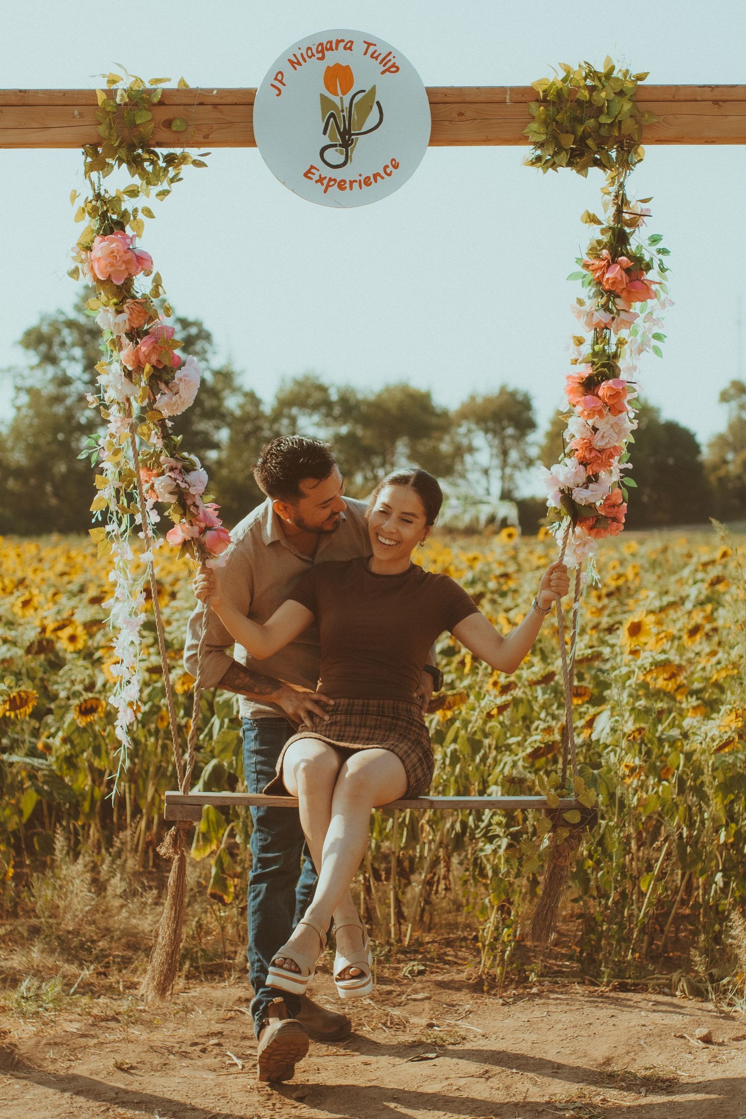 Two people share a loving moment among sunflowers on a sunny day with floral arch decorations.