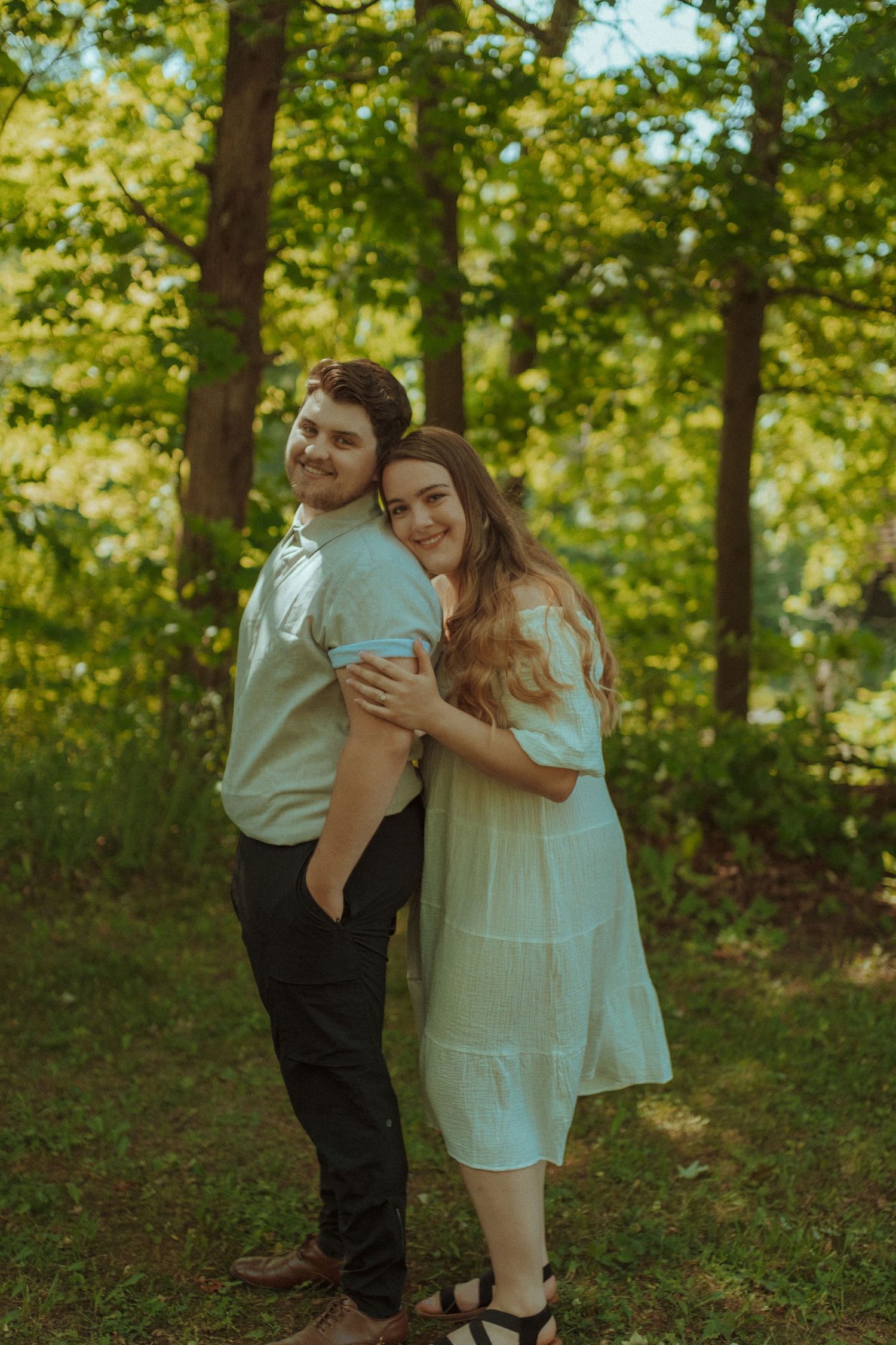 Two people share a sweet embrace while standing together in a sunlit forest with lush green foliage in the background.