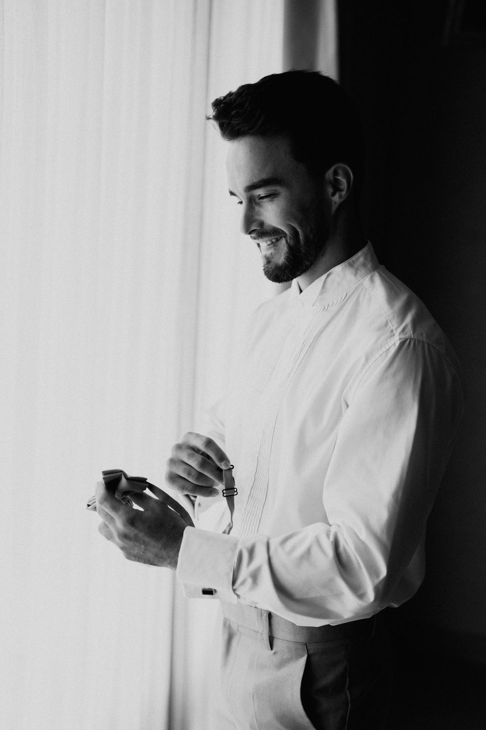Black and white portrait of someone adjusting formal attire near a window.