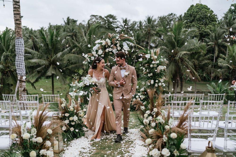 Tropical outdoor wedding ceremony setup with white chairs and floral arrangements under palm trees.