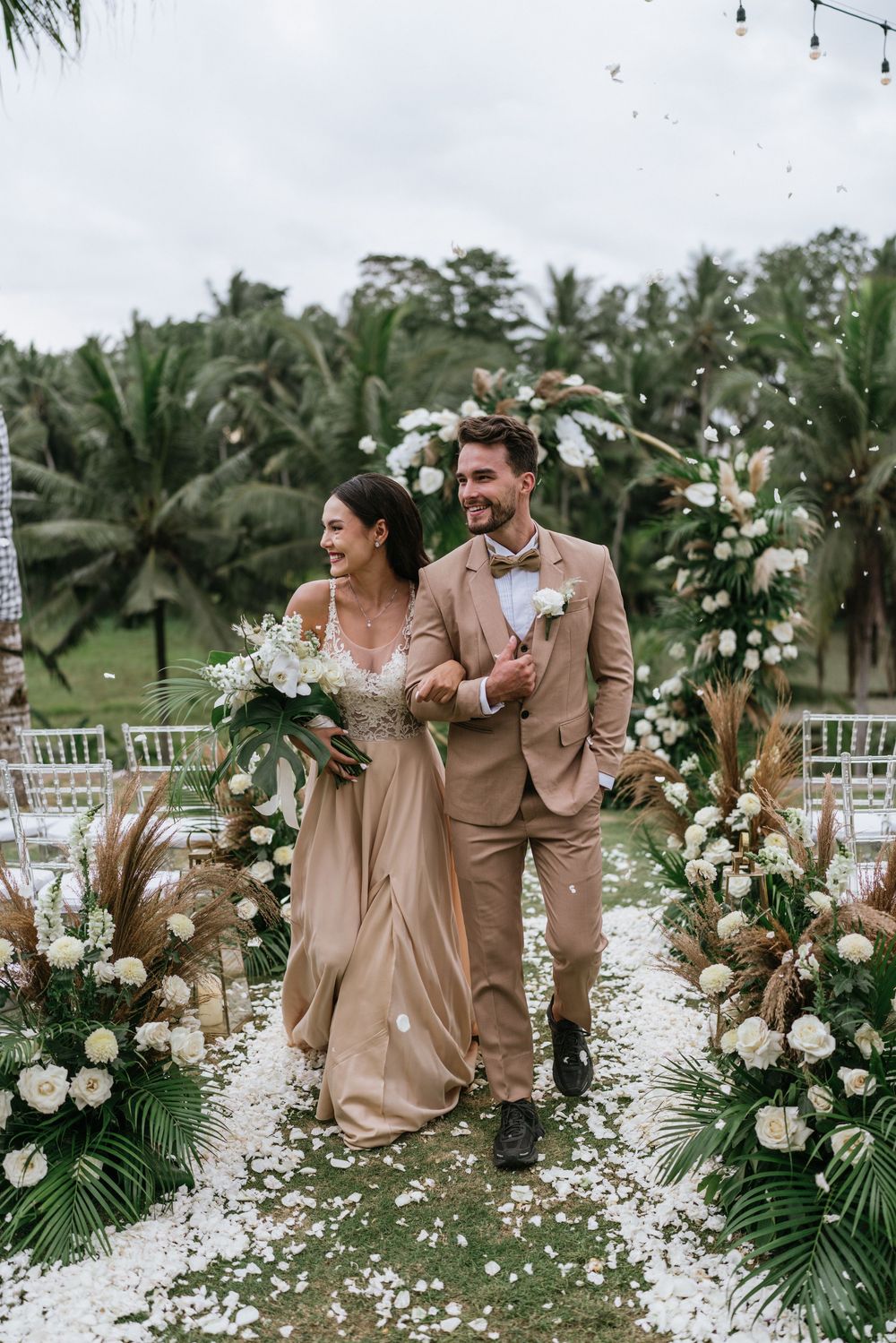 Wedding party in neutral tones walking through a tropical garden setting with floral arch decorations.