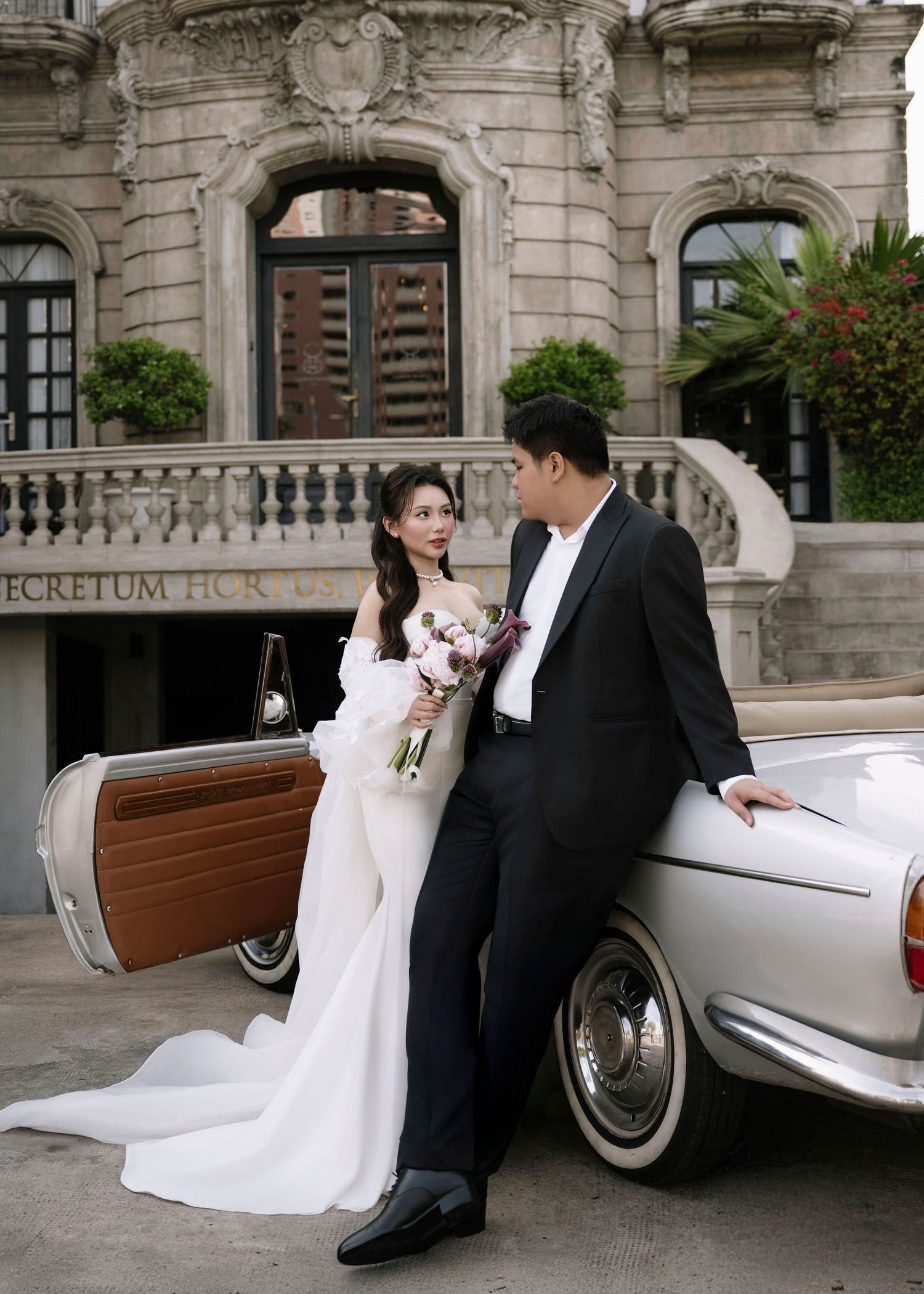 Elegant wedding couple pose beside a classic white vintage car outside a grand ornate building.