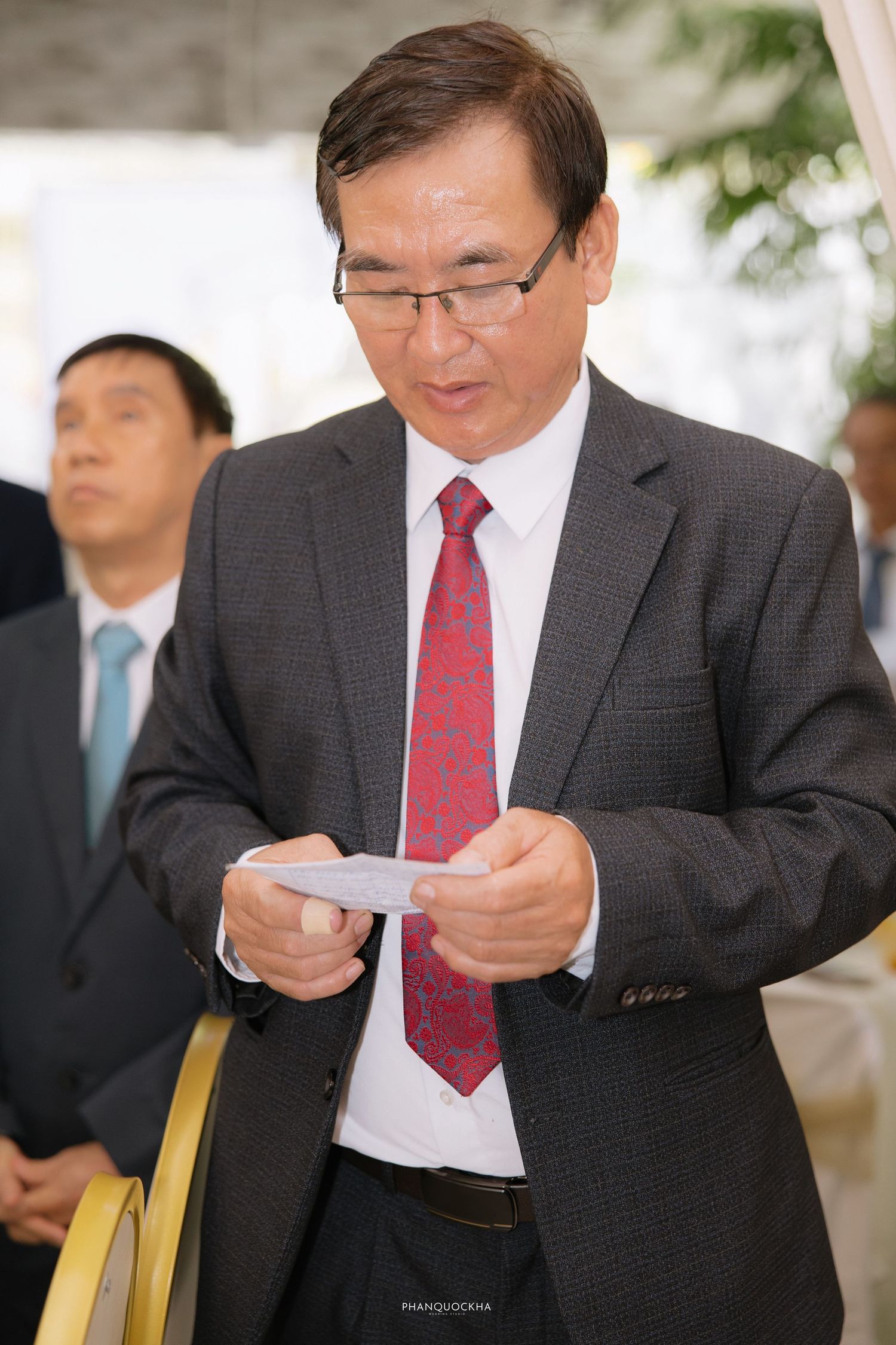 Asian man in gray suit and red tie reads a document at a formal wedding event.