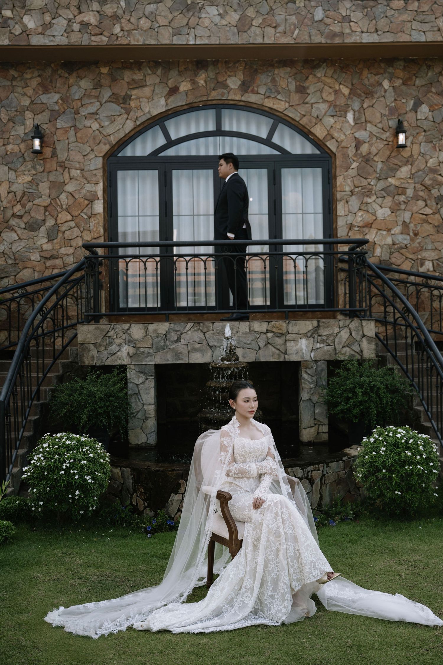 Bride in white lace gown seated outdoors while groom stands on balcony above, in front of a stone villa.